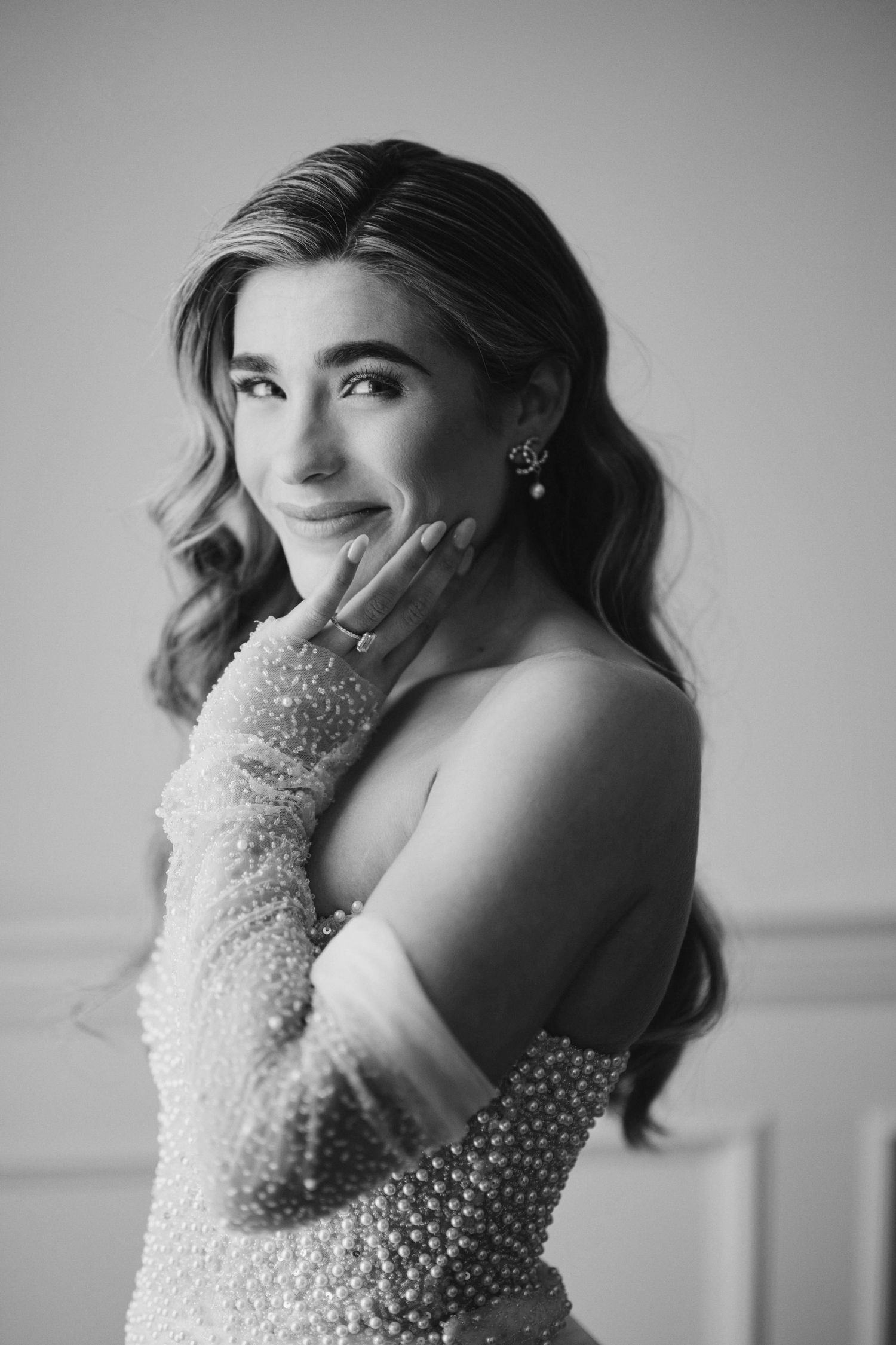 Black and white portrait of elegant woman in pearl dress and gloves, smiling gracefully with hand near face.