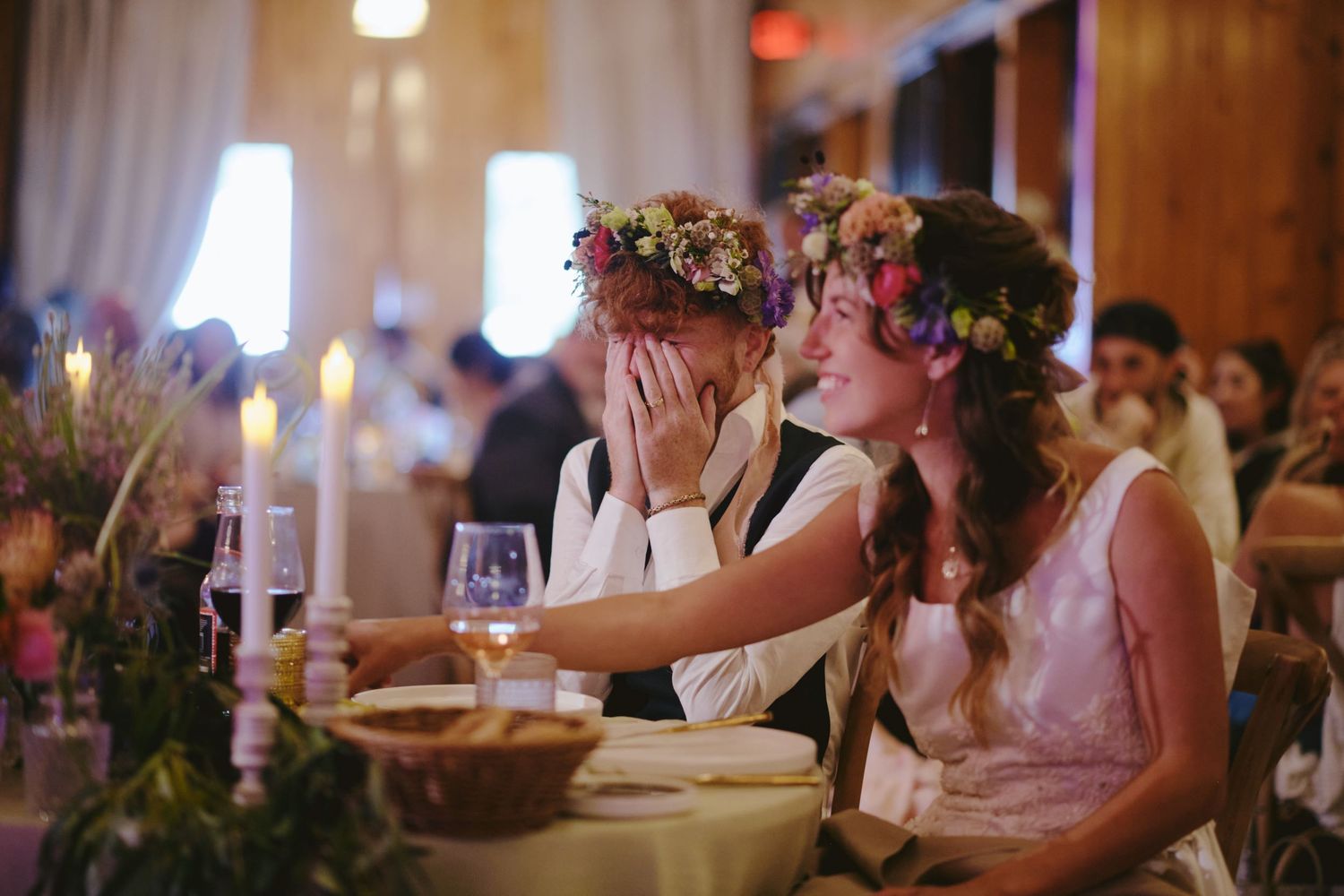 Two women wearing floral crowns share an emotional moment at a wedding reception dinner table with candles.