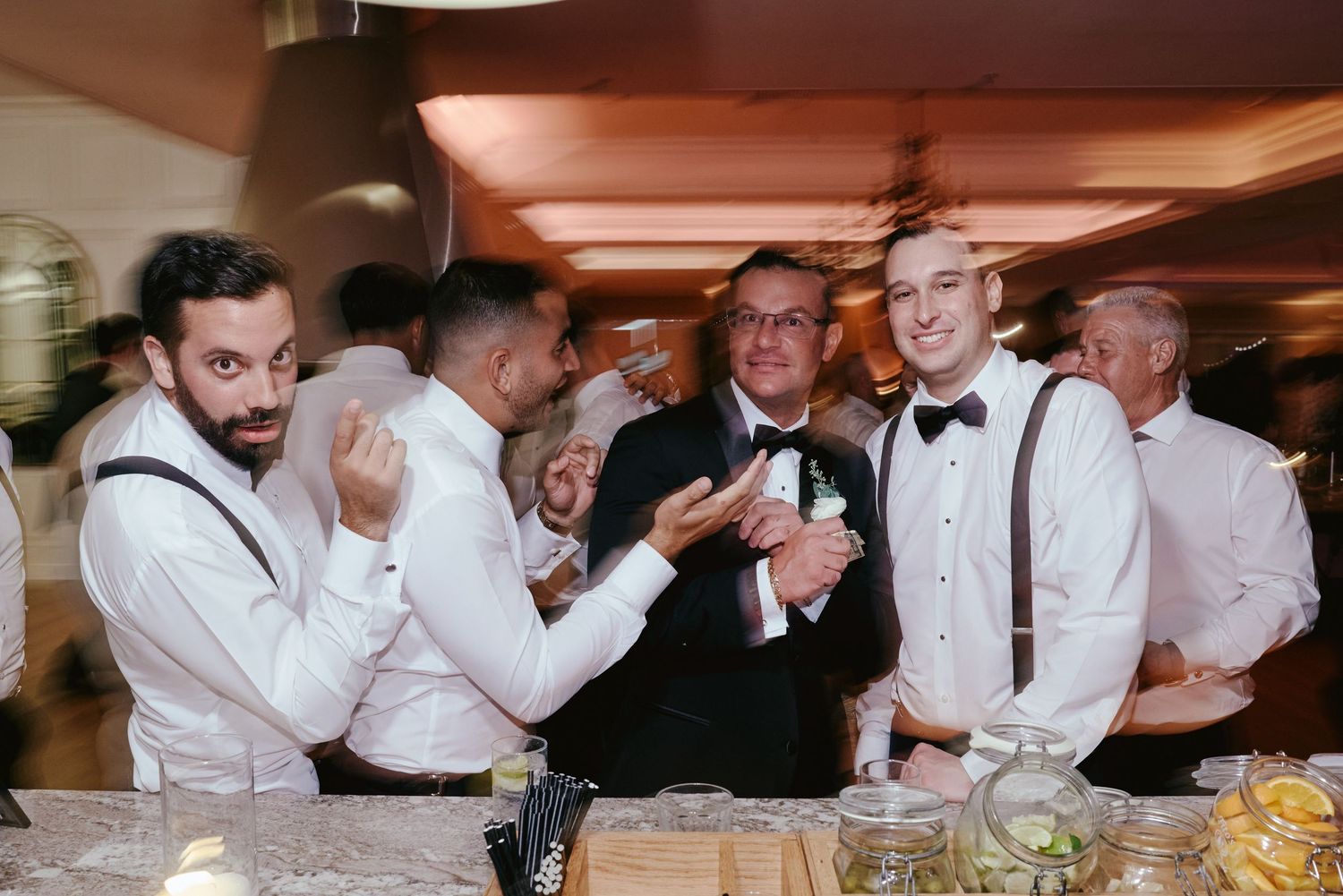 Bartenders and waitstaff dressed in formal attire share a lively moment behind the bar at an elegant event.