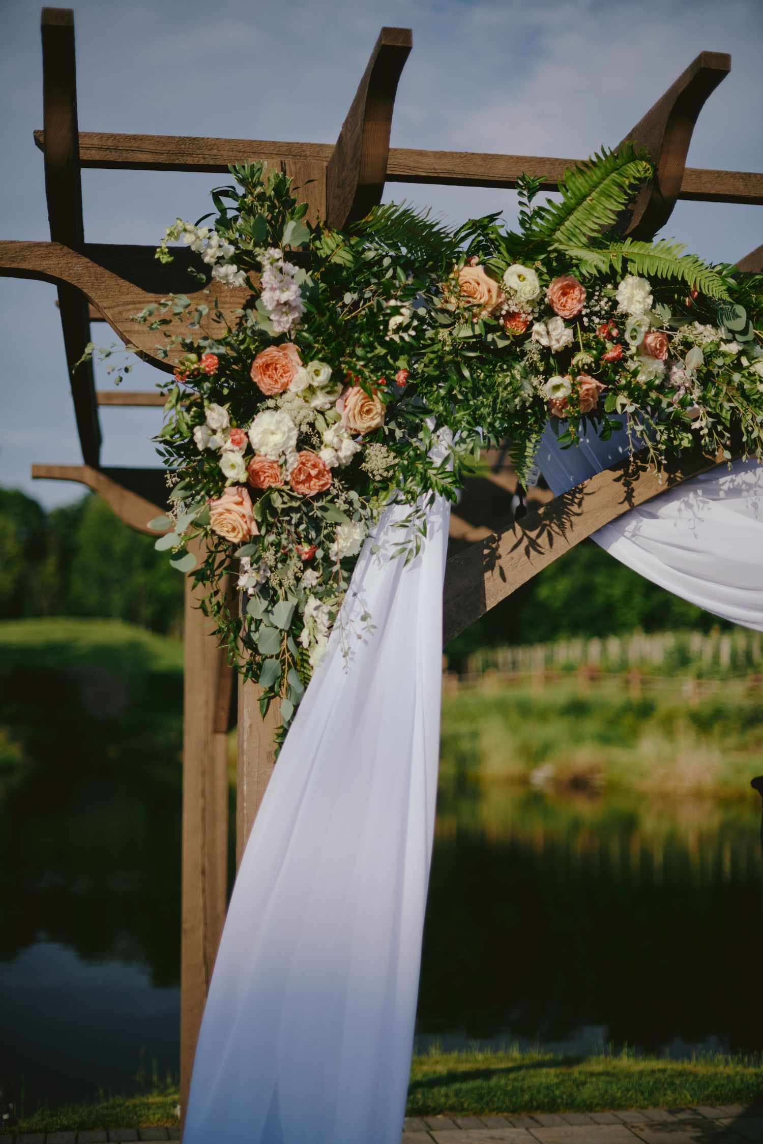 Wooden wedding arch decorated with peach roses, white flowers and greenery against lake backdrop with white fabric draping.