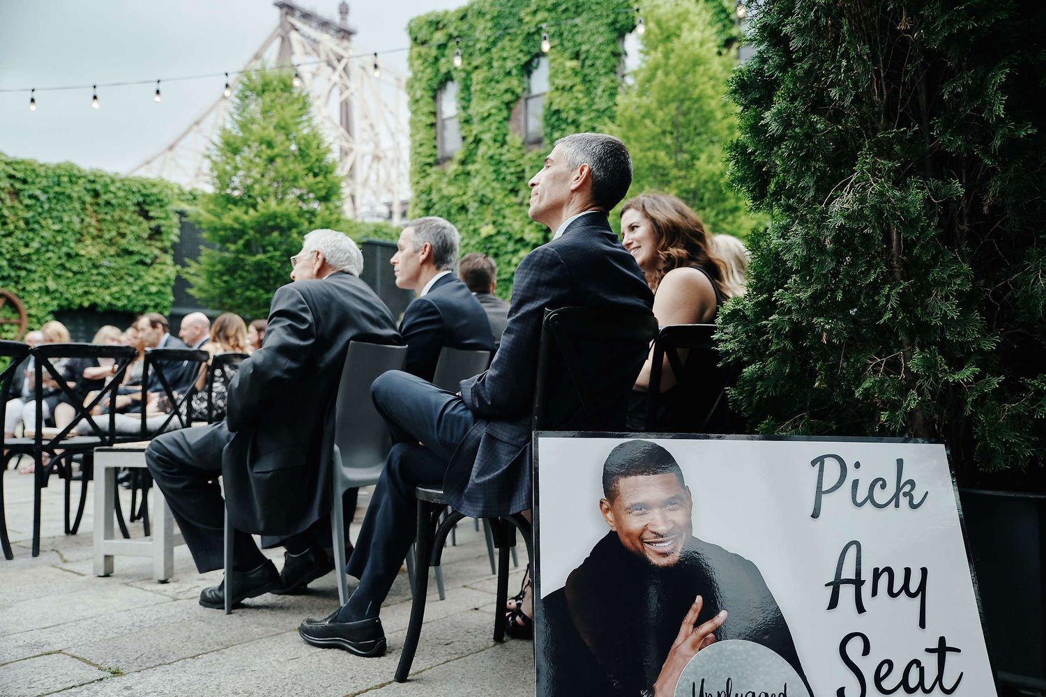 Wedding guests sit at outdoor tables while a pick-any-seat sign is displayed during a casual outdoor celebration.