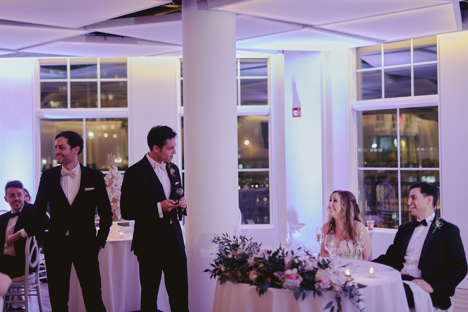 Wedding party gathers in a modern white venue with purple lighting and large windows during an evening reception.