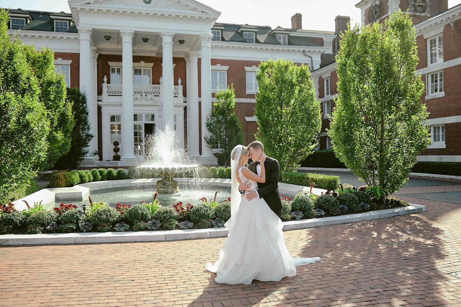 A couple shares an embrace in front of a grand white-columned building with manicured gardens and brick pathways.