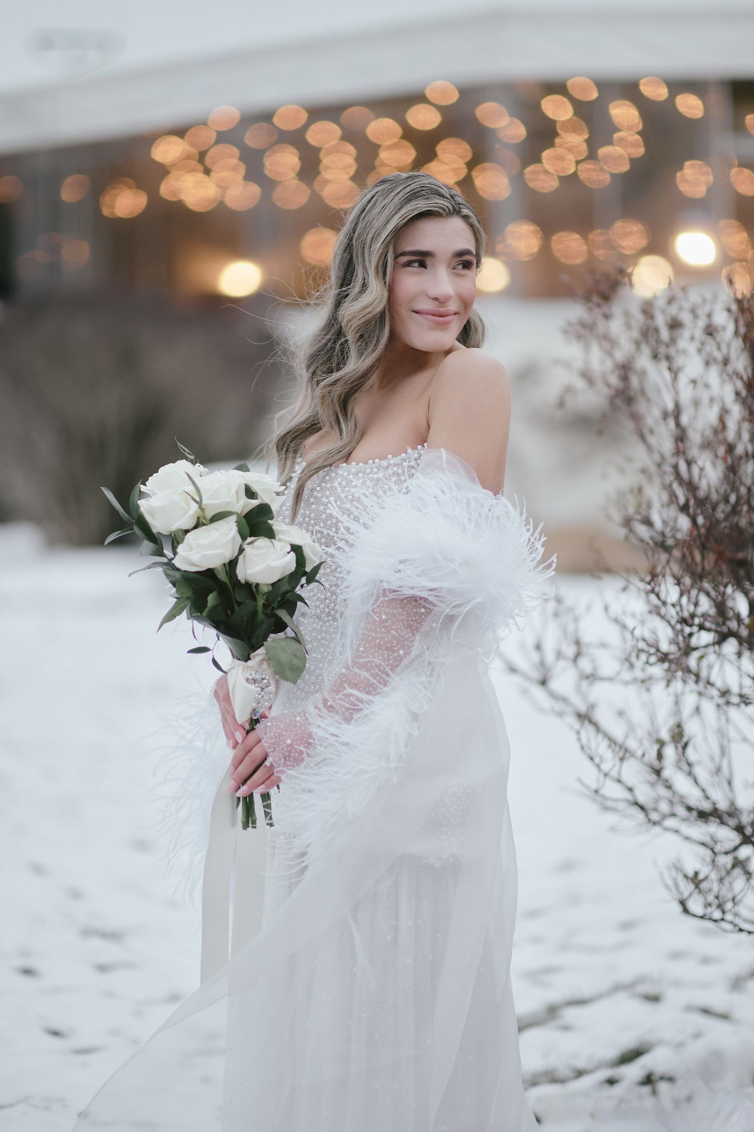 A bride in an off-shoulder lace gown holds white roses against a snowy winter backdrop with twinkling bokeh lights.