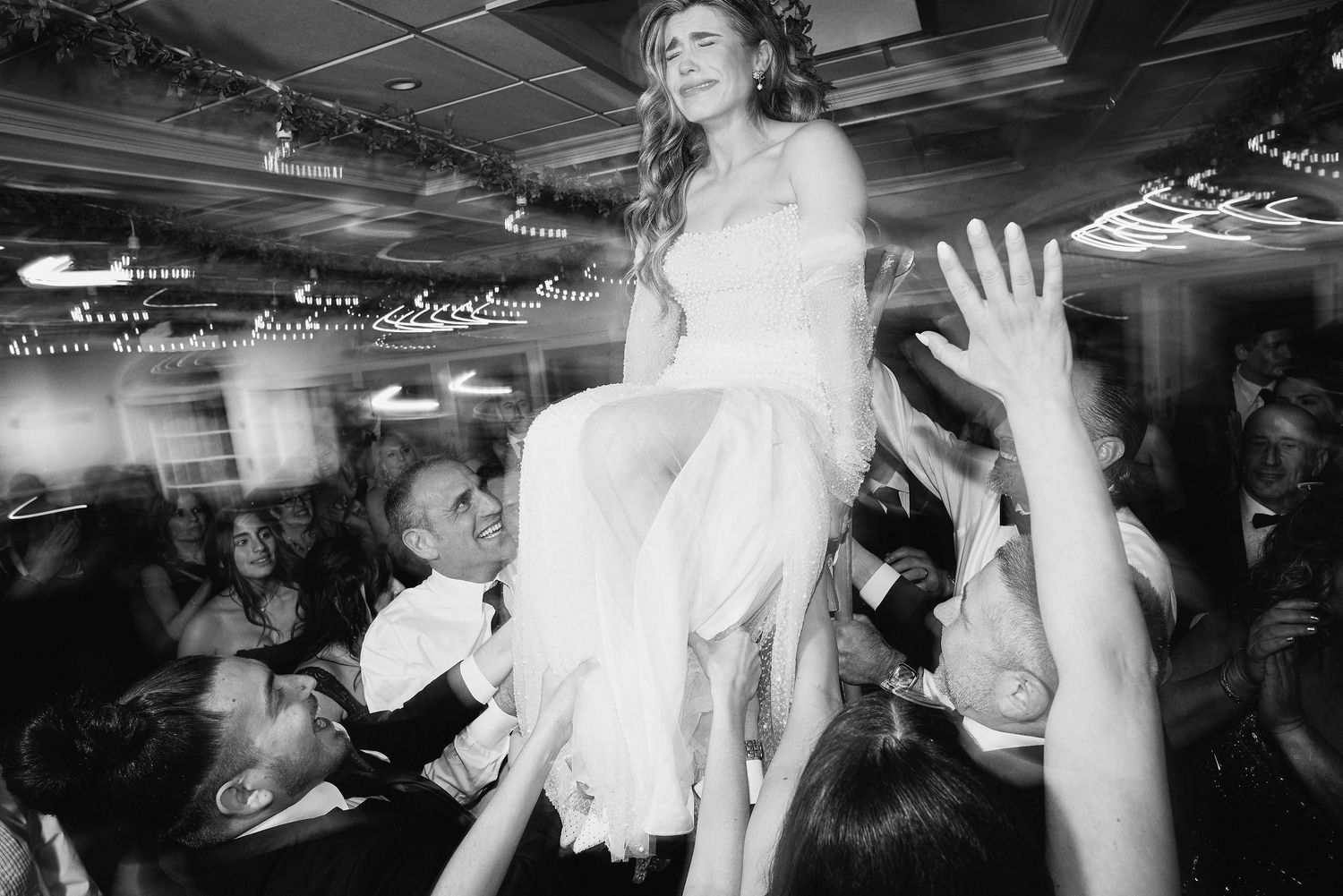 Bride being lifted up during wedding reception celebration while guests reach up with raised hands.
