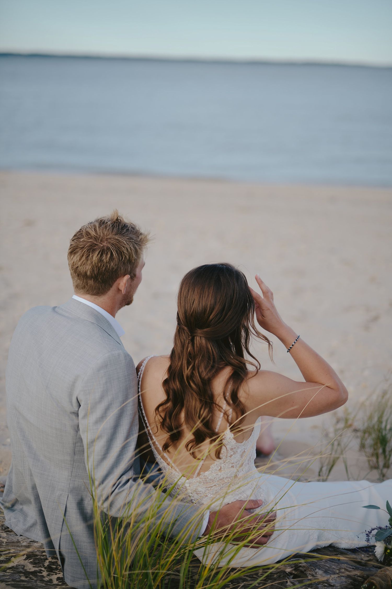 A couple sits together on a beach blanket at sunset, looking out over calm waters through coastal grass.
