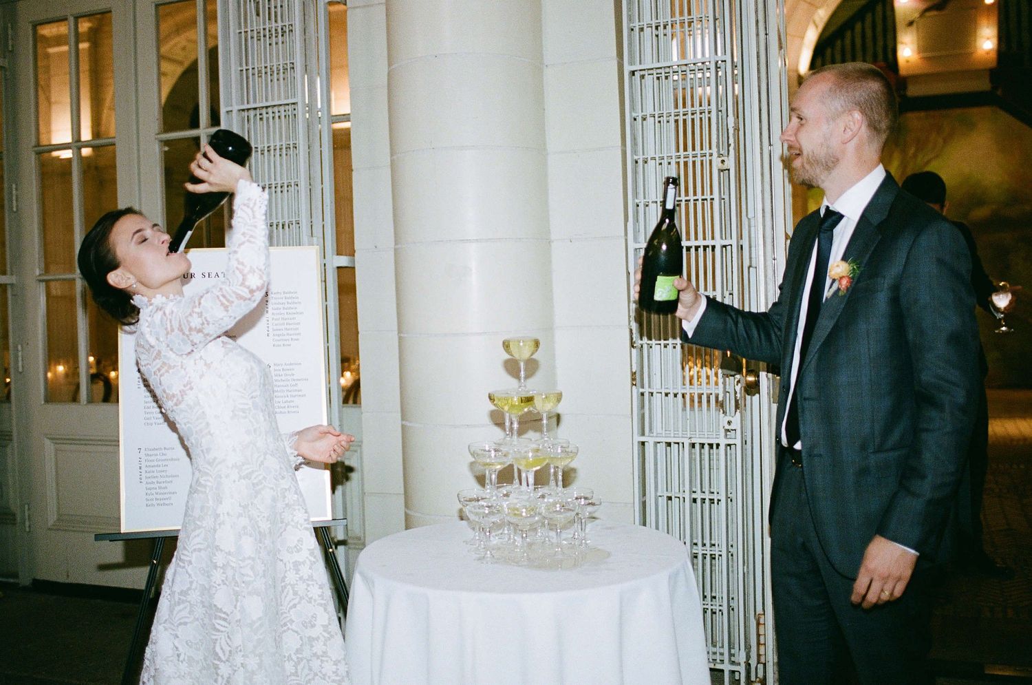 Two people in formal attire playfully toast with champagne bottles beside an elegant white wedding cake.