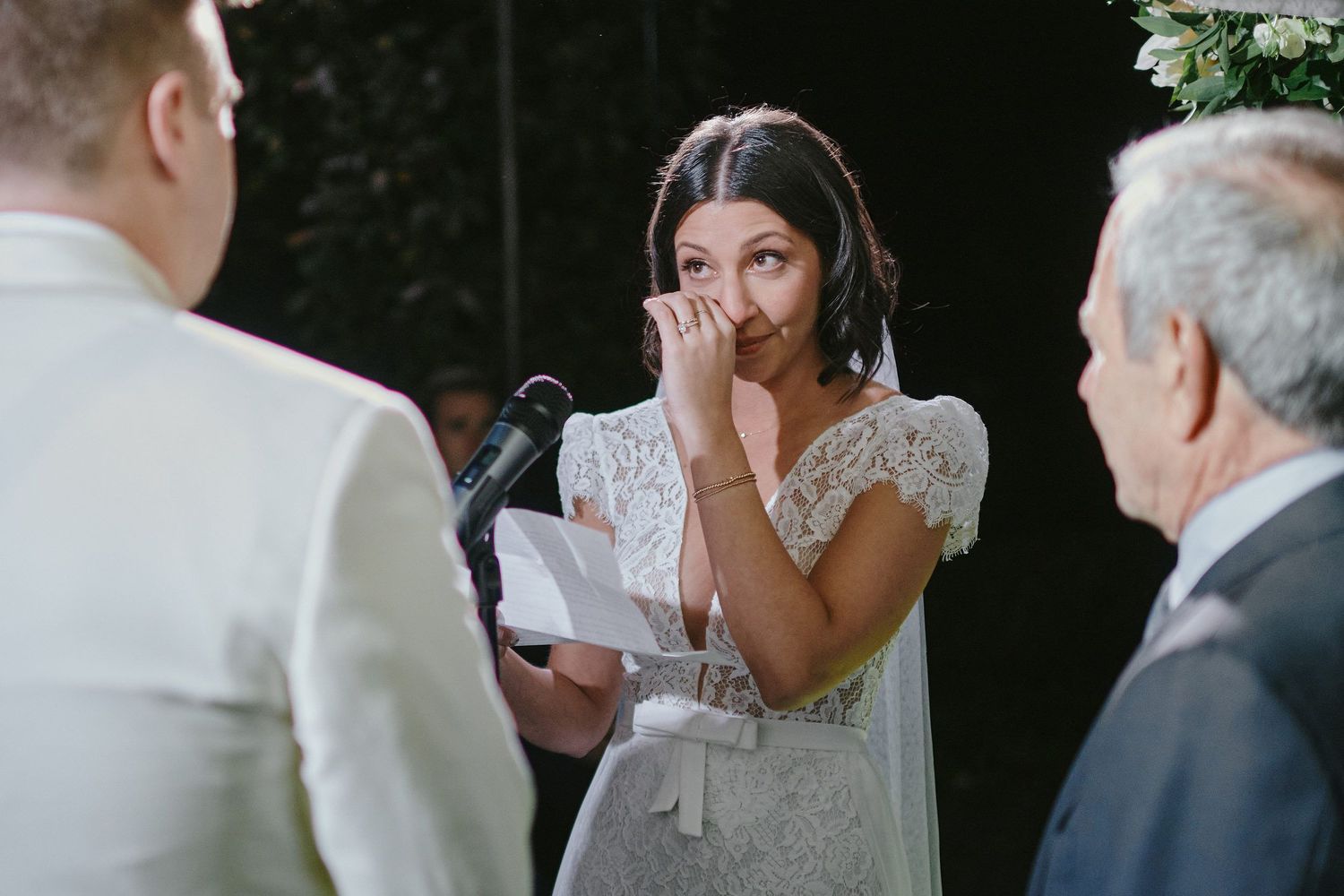 A bride in a lace wedding dress becomes emotional during an outdoor ceremony while reading vows.