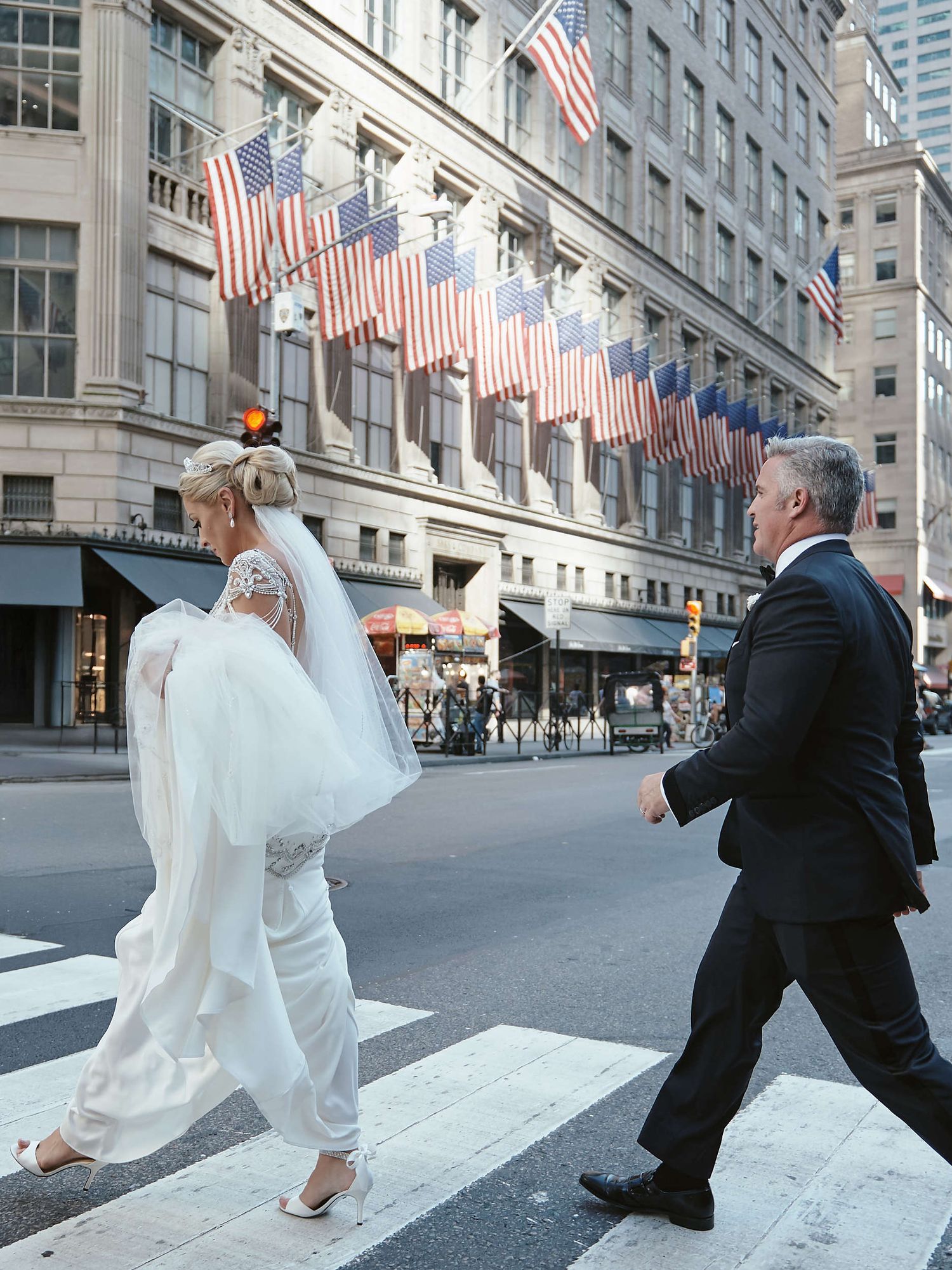 A couple crosses a city street on their wedding day with American flags displayed on the building behind them.
