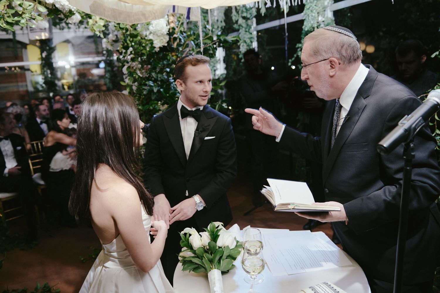 A wedding ceremony takes place at night under twinkling lights with an officiant speaking to the couple at the altar.