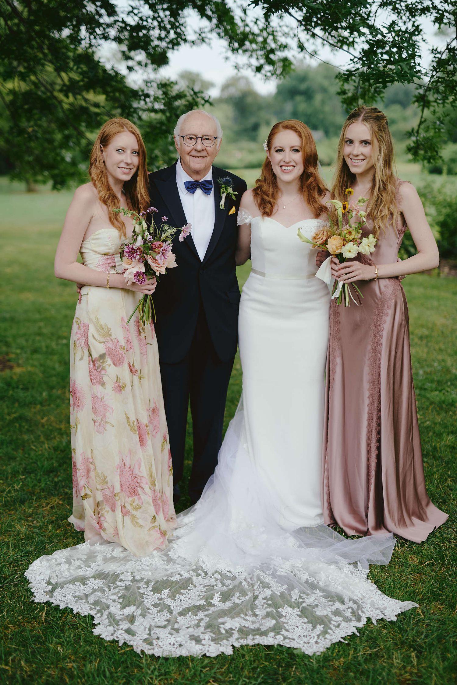 Wedding party of four people standing outdoors on grass with beautiful floral arrangements and elegant formal attire.