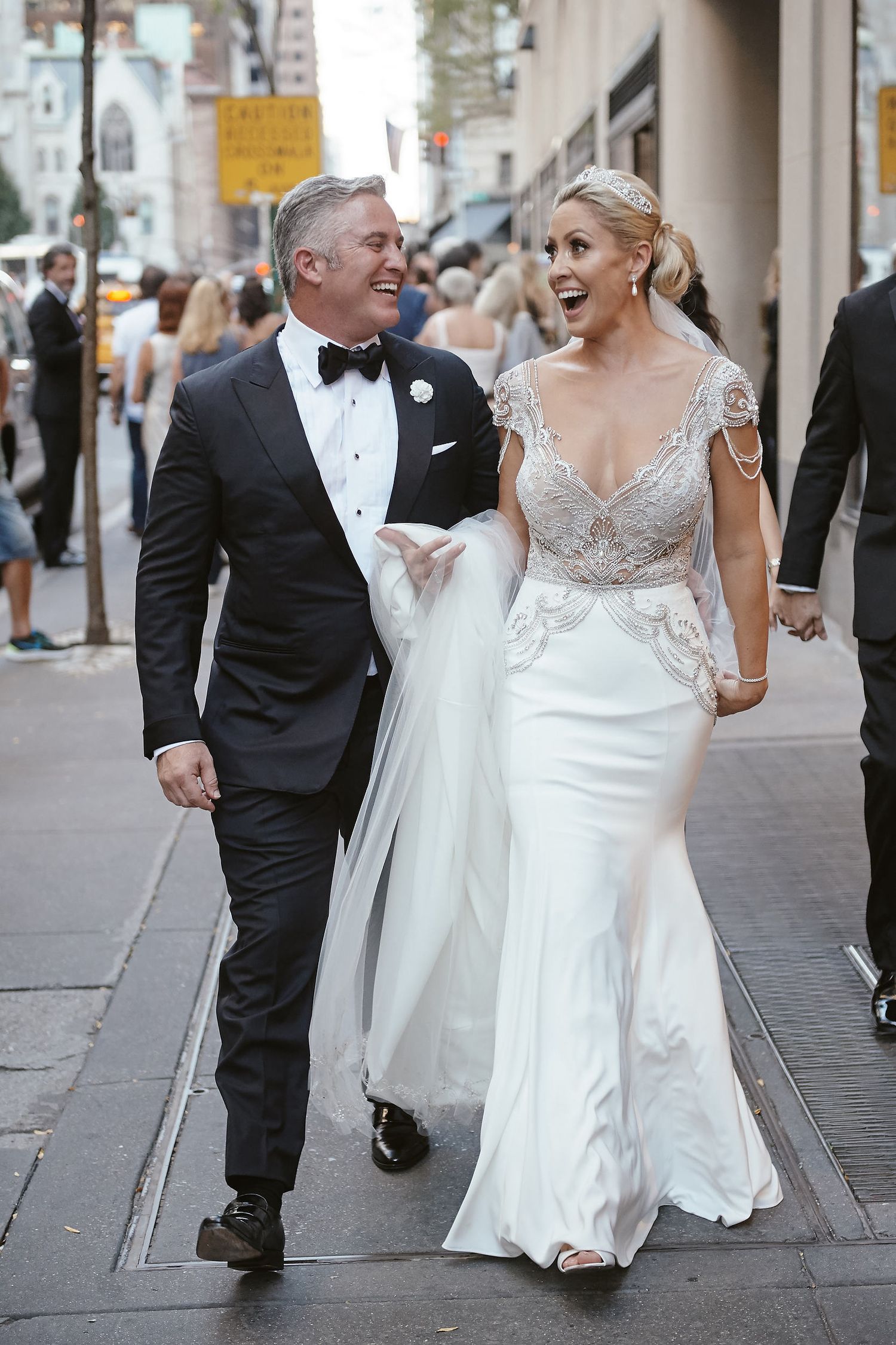 A bride in a lace-detailed fitted gown walks down a city street with her groom wearing a classic black tuxedo and bowtie.