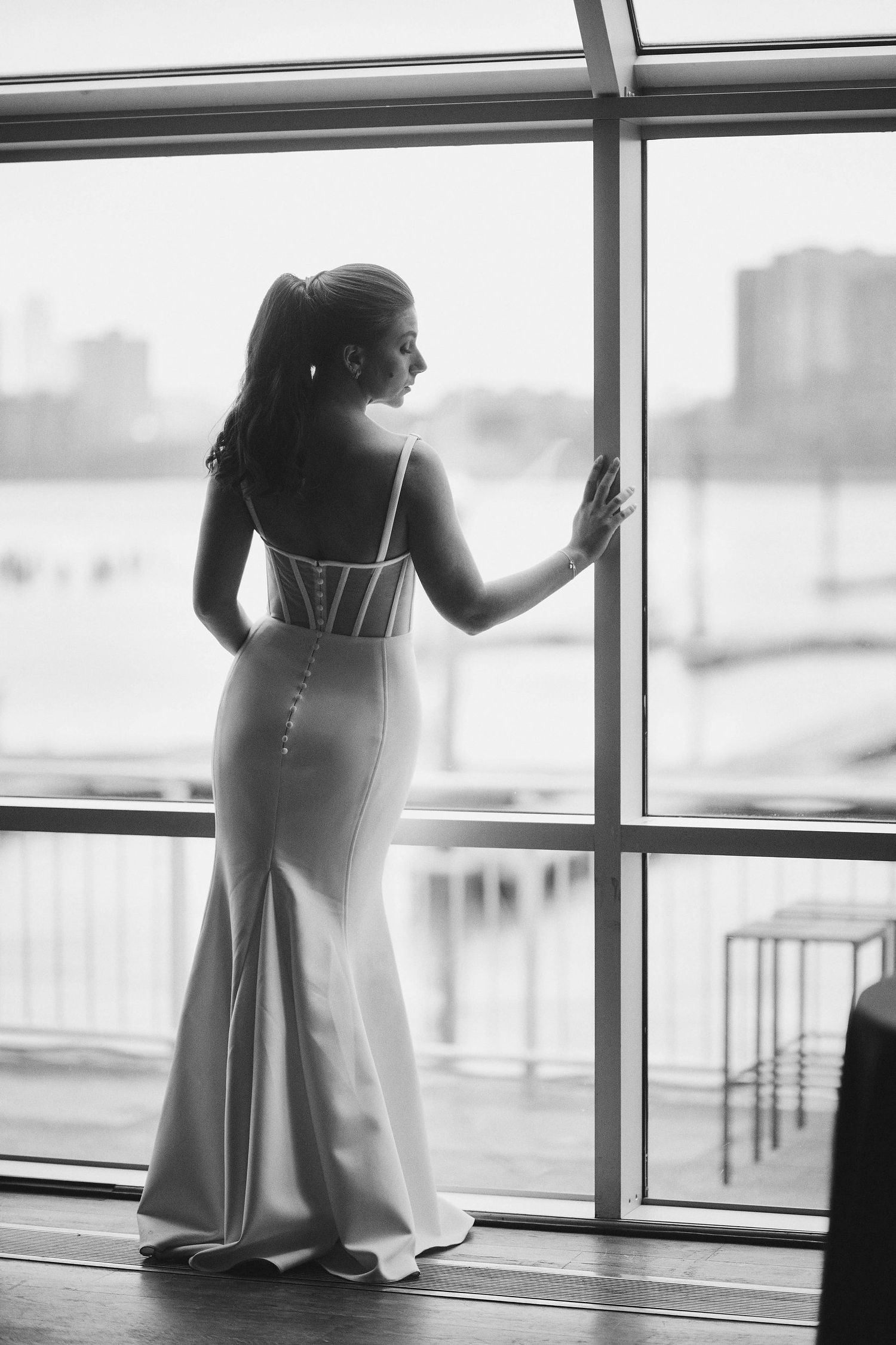 A silhouetted figure in an elegant fitted dress stands gazing through large windows at a cityscape in black and white.