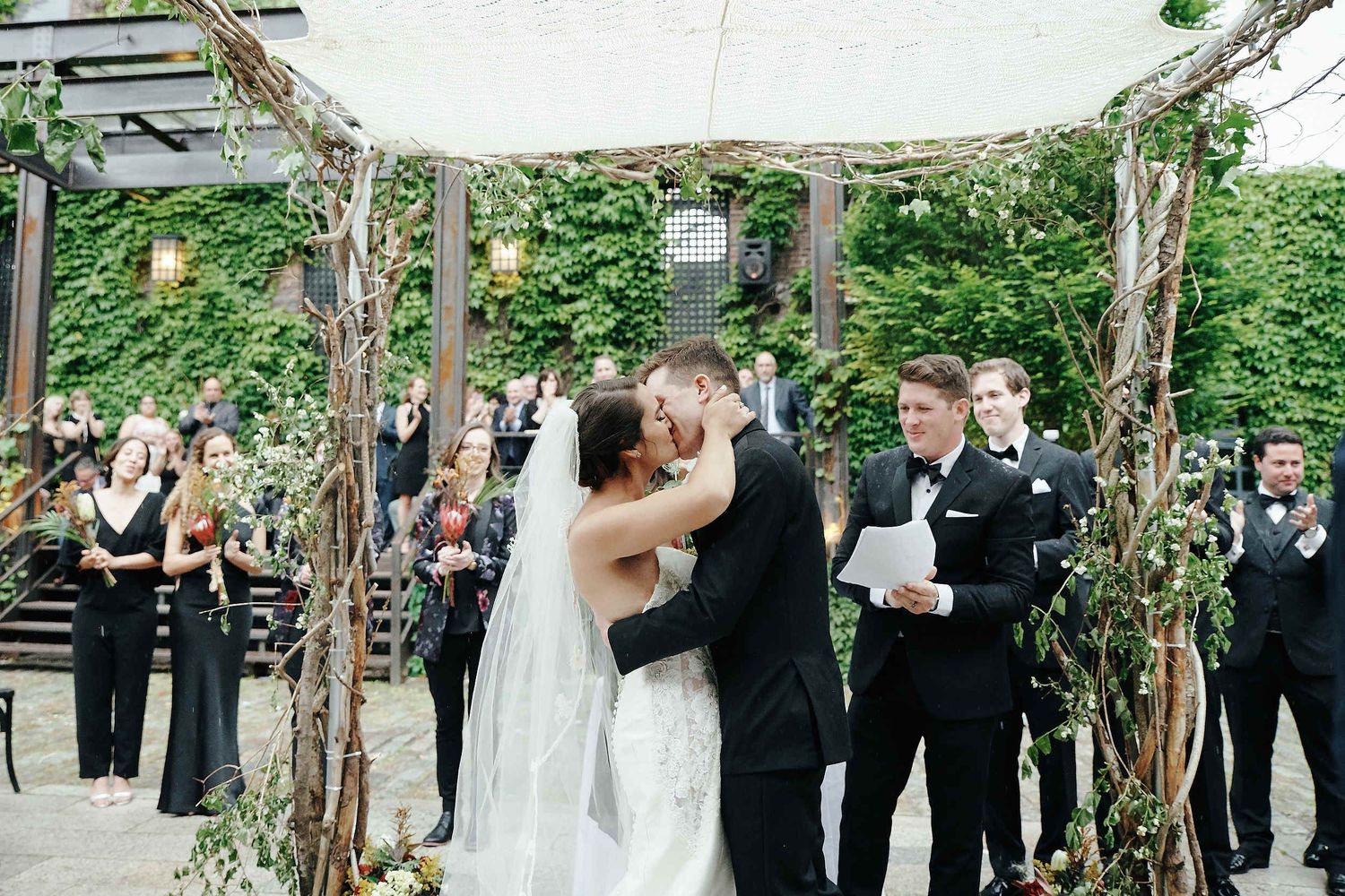 A wedding ceremony takes place under a rustic wooden pergola decorated with greenery and string lights.