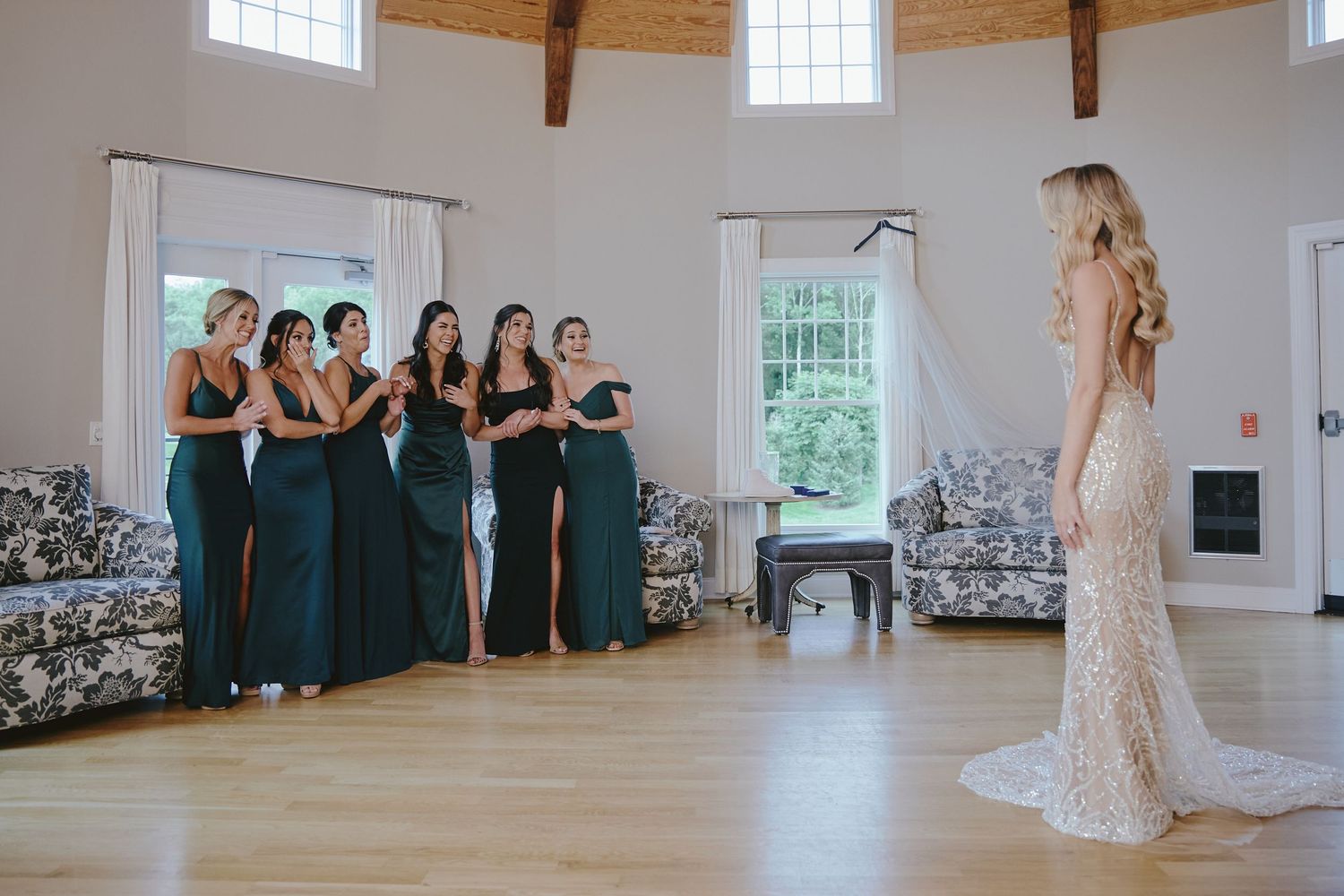 A bride in a lace gown stands before her bridesmaids wearing teal dresses in a bright white room with wooden floors.