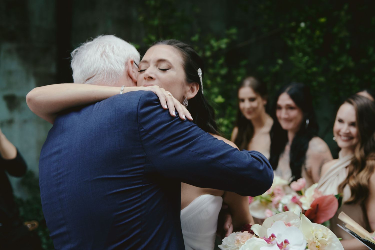 Two people embrace at an outdoor wedding ceremony while guests look on with smiles.