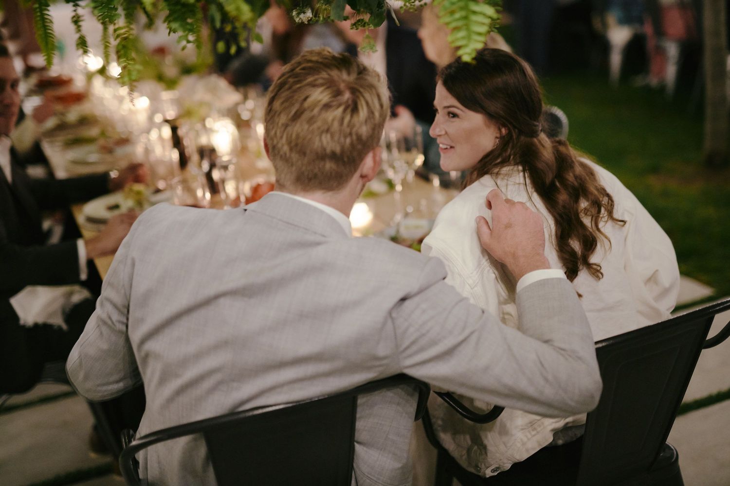 A couple sharing an intimate moment at a candlelit outdoor dinner table surrounded by greenery.