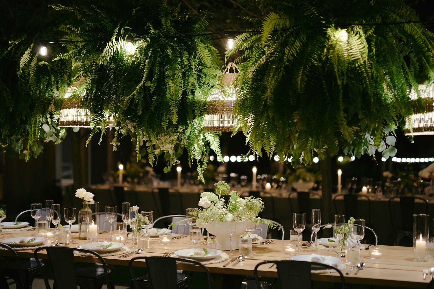Elegant table settings beneath hanging ferns and warm lighting at an indoor reception venue.