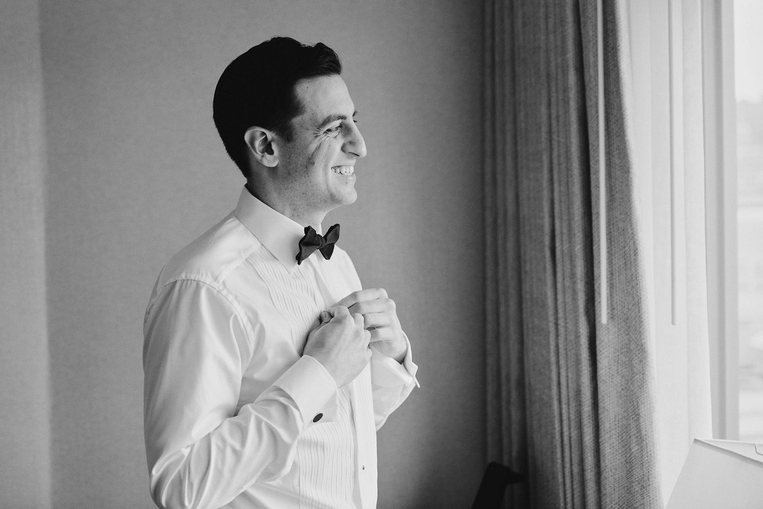 Black and white photograph of a person adjusting their bowtie while getting ready near a window.