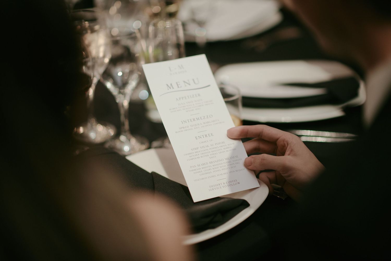 Hand holding an elegant restaurant menu card at a table setting with white plates and wine glasses in soft lighting.