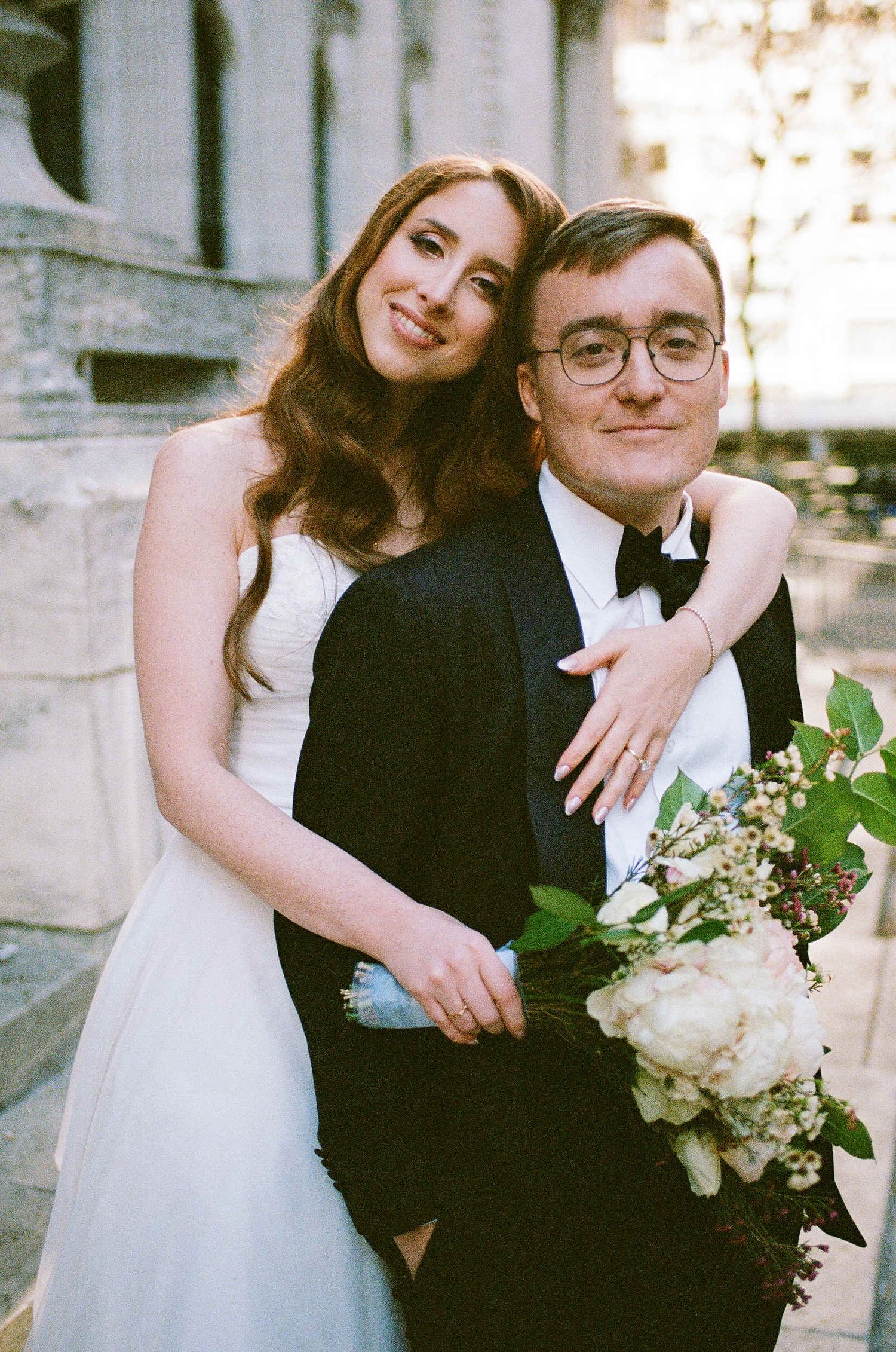A bride in white wedding dress holding flowers embraces groom in black tuxedo and bowtie during outdoor wedding ceremony.