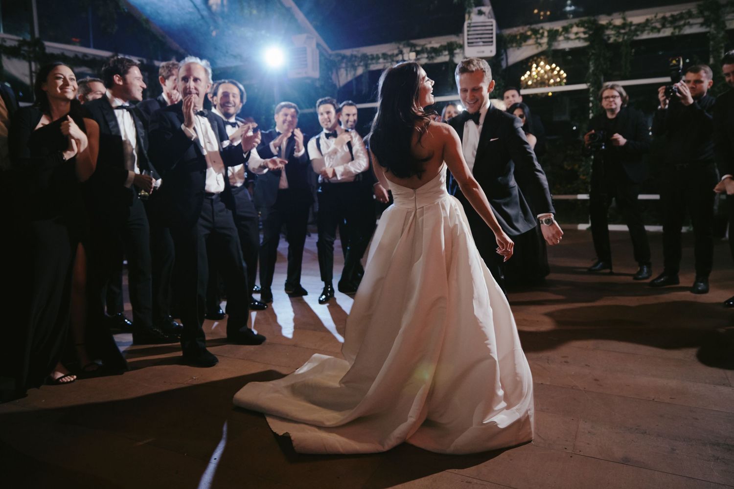 A person in a flowing white ball gown dances on a dark dance floor while guests watch in formal attire.