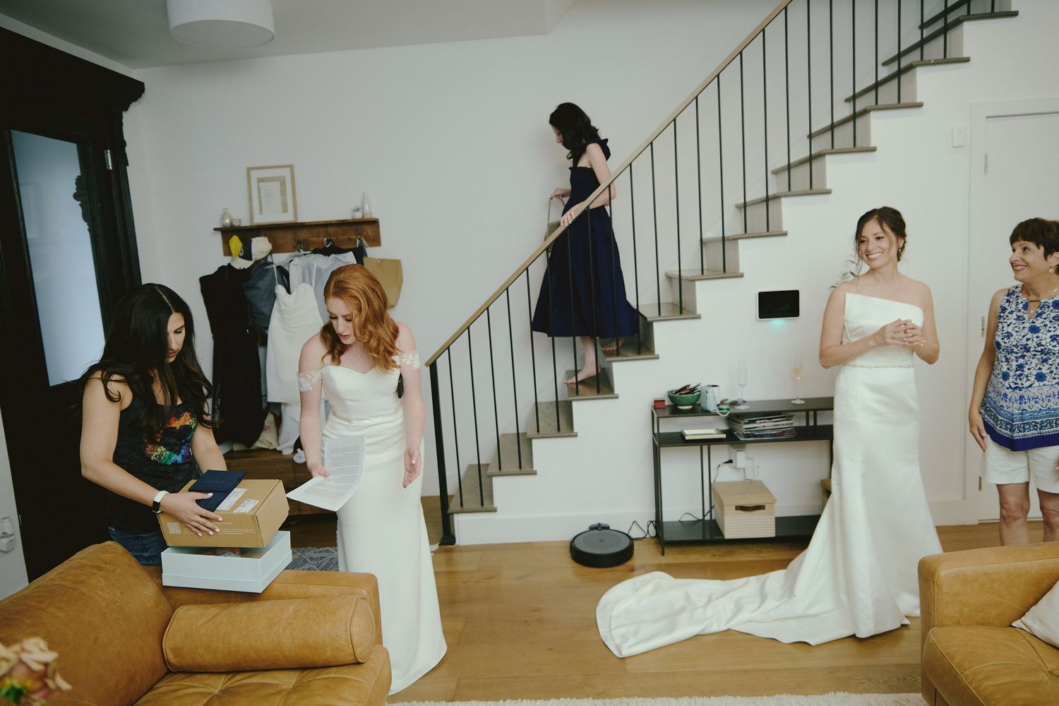 A group of people gathered in a bright living room with white walls and a staircase, with formal attire and gowns.