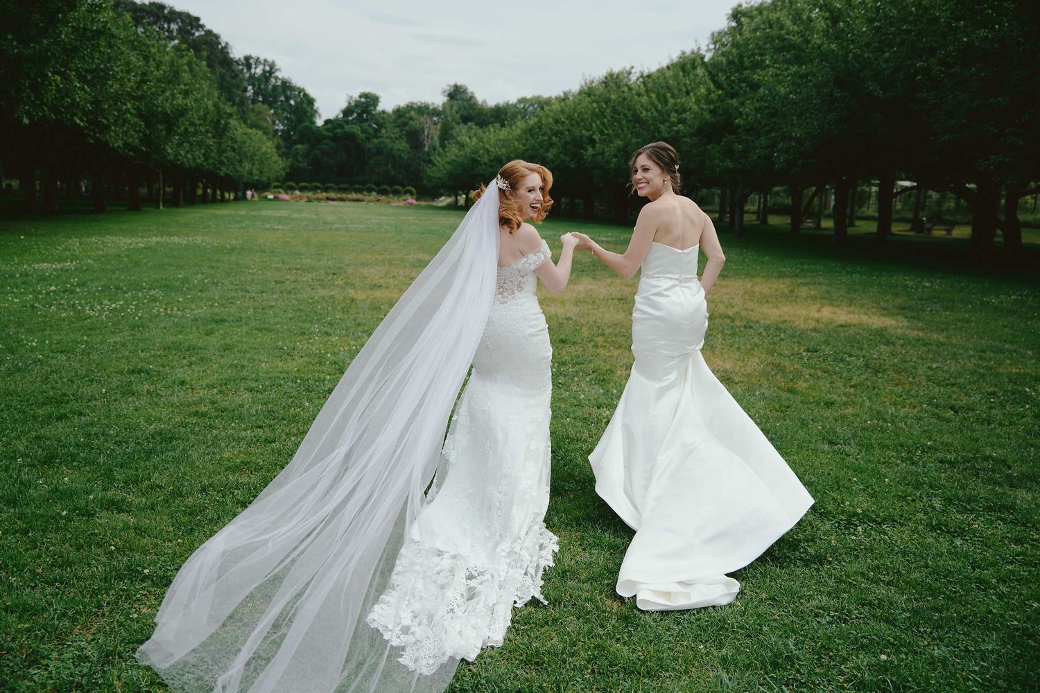 Two brides in elegant white mermaid wedding dresses share a joyful moment on a lush green lawn with trees in background.