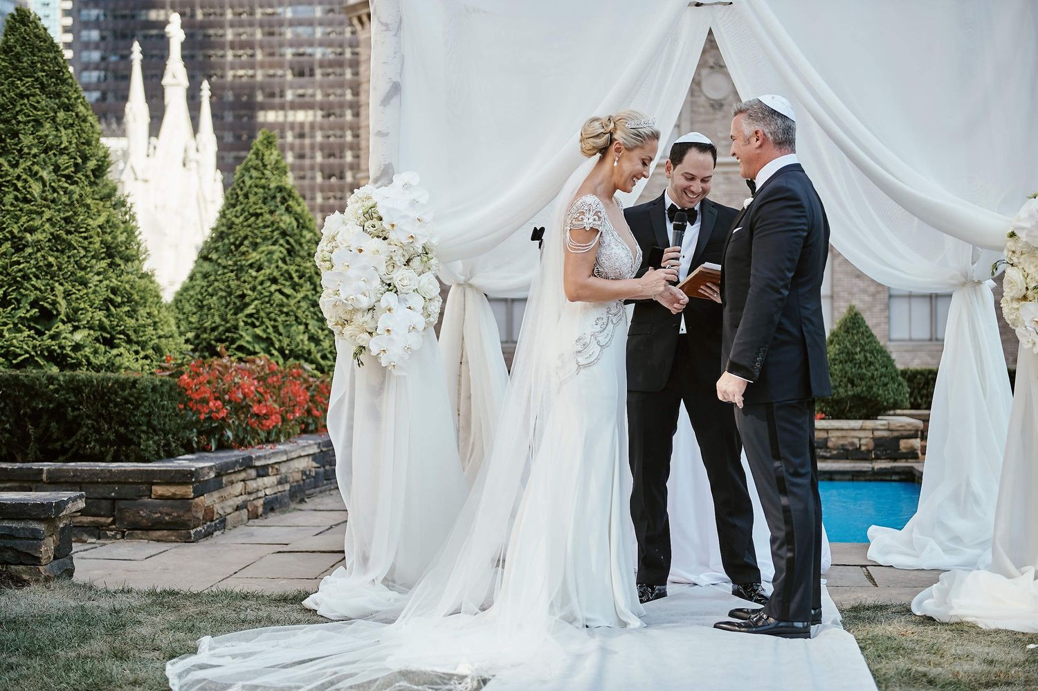 An elegant outdoor wedding ceremony takes place on a rooftop terrace with white drapes and floral decorations.