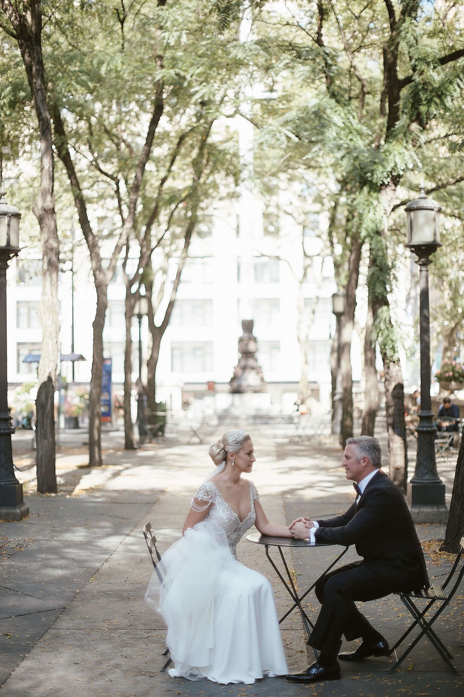 A couple sits on bistro chairs in a tree-lined park pathway with a fountain visible in the background.