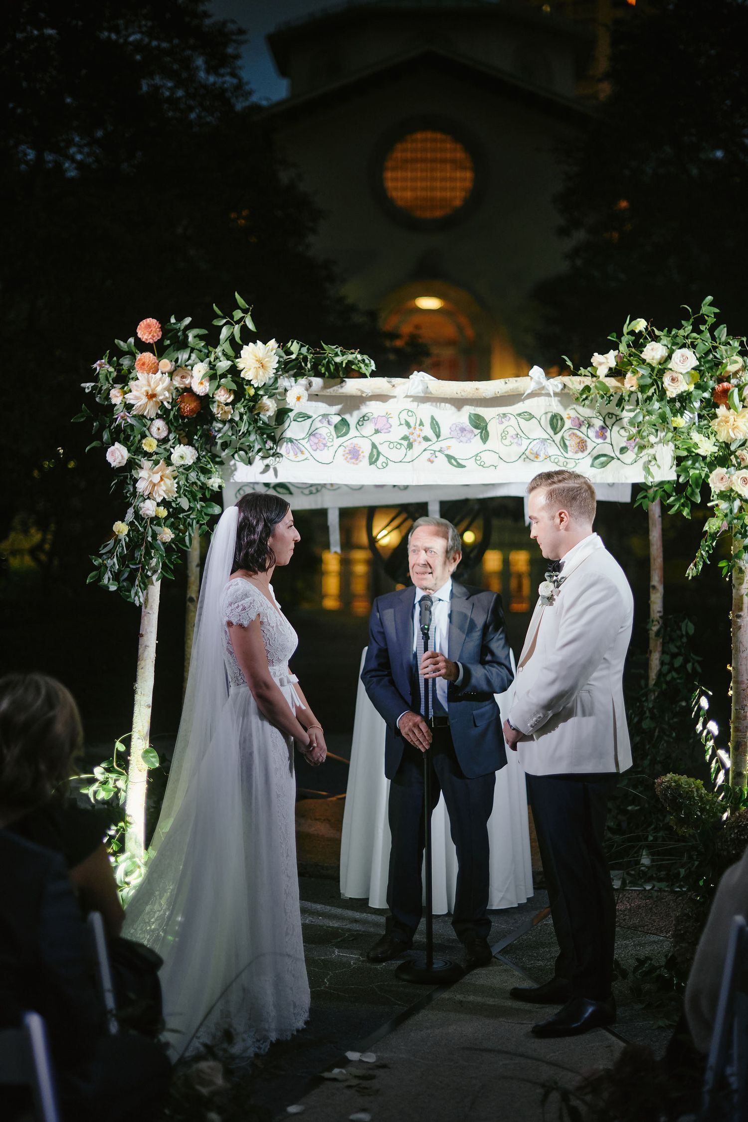 A romantic outdoor wedding ceremony under a floral-adorned white arch at dusk with soft lighting creating an ethereal atmosphere.