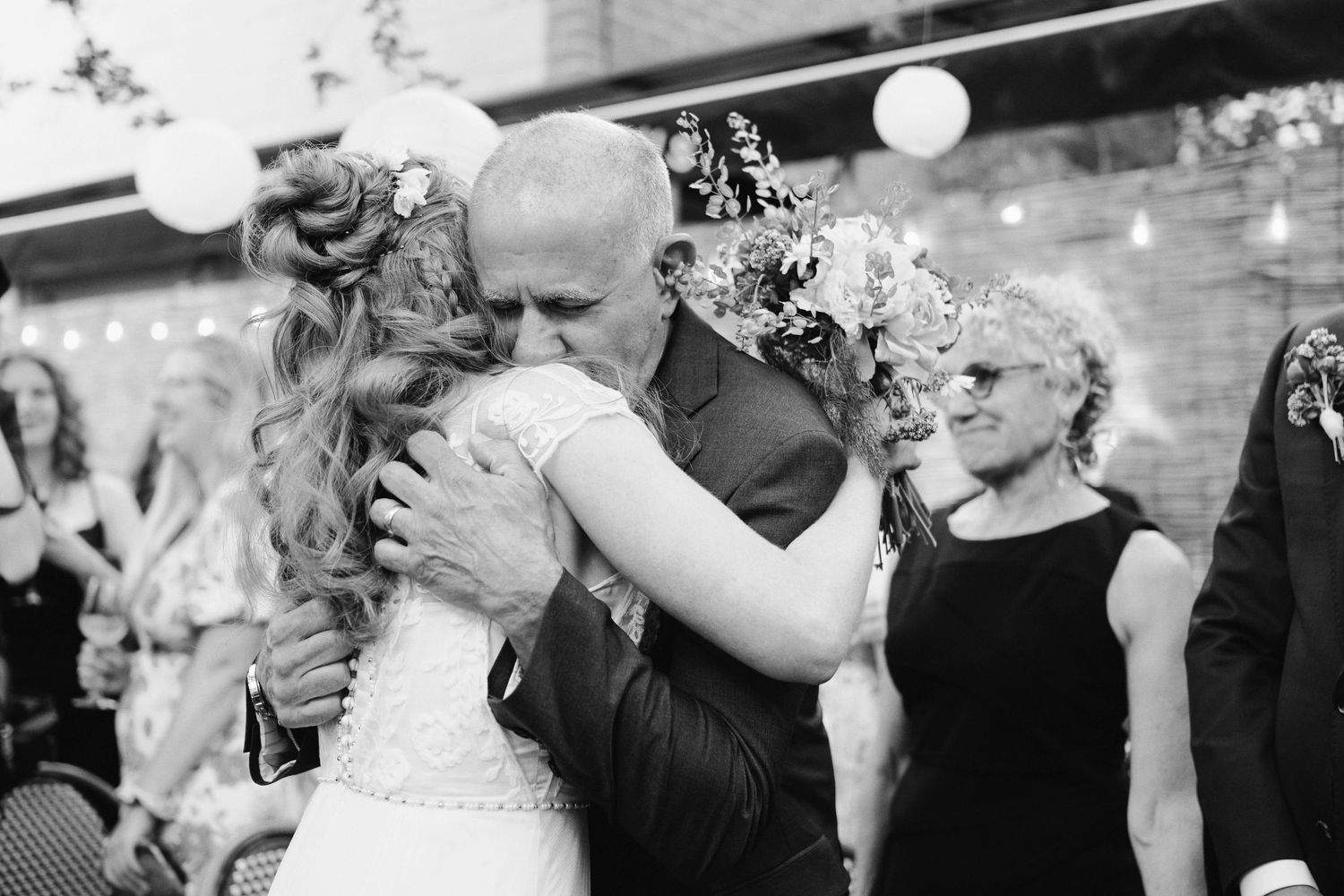 Black and white photo of a bride sharing an emotional hug with a bald man at an outdoor wedding reception.