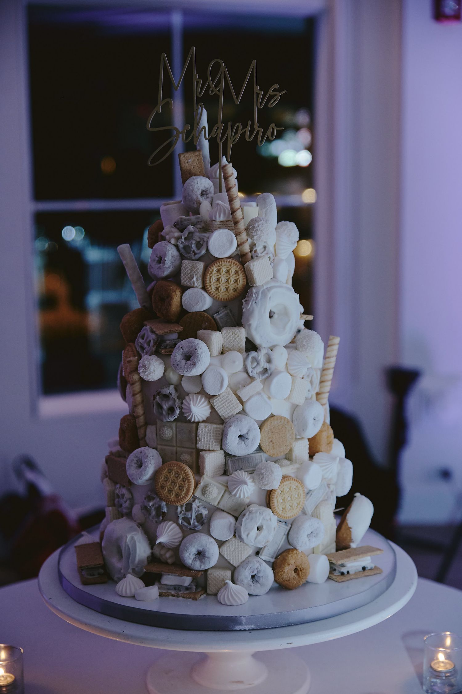 A tiered wedding cake decorated with an assorted mix of white and beige cookies and macarons on a white cake stand.