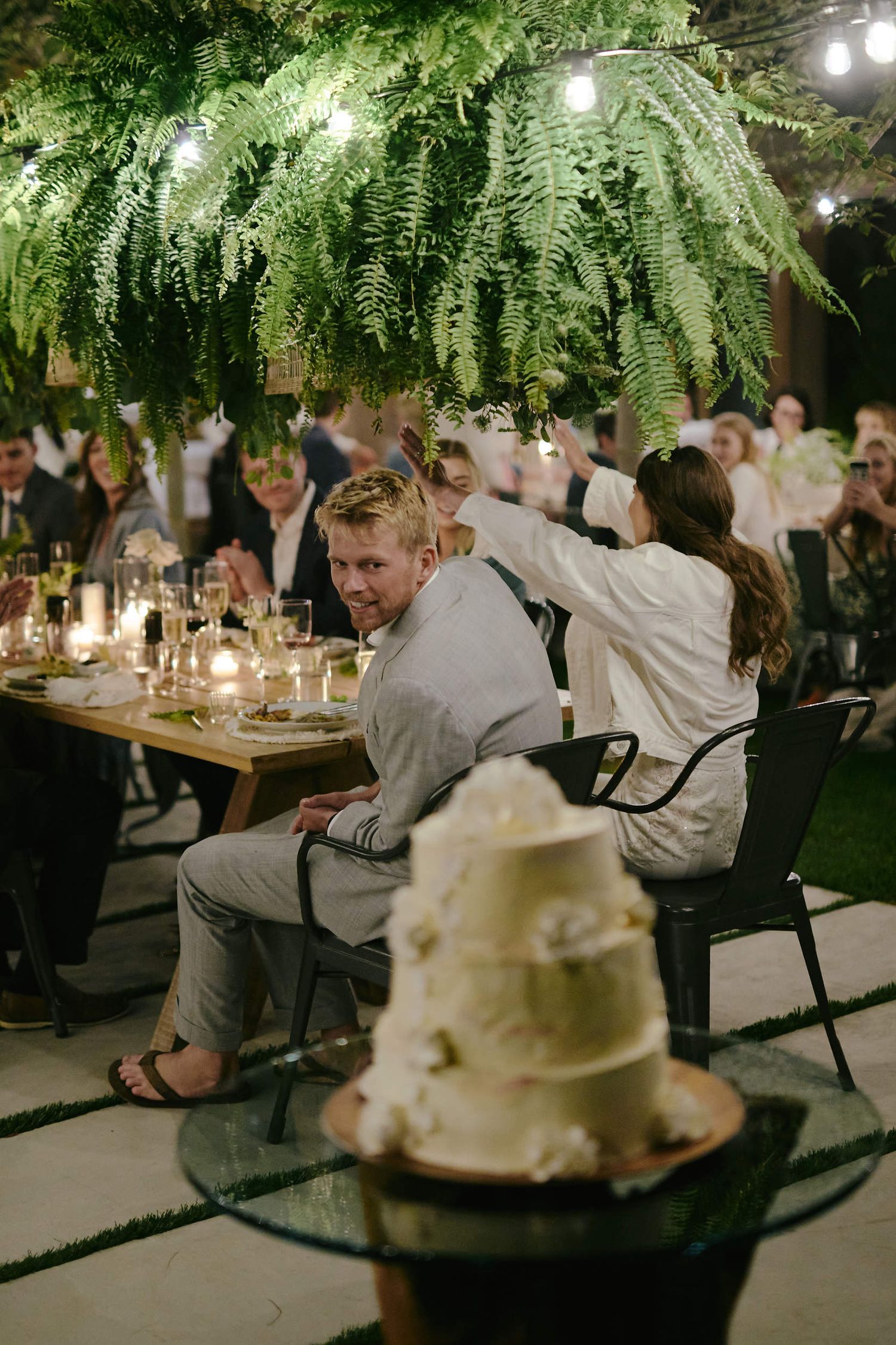 Evening wedding reception with hanging ferns, glowing lights, and tiered white cake on display as guests dine at tables.
