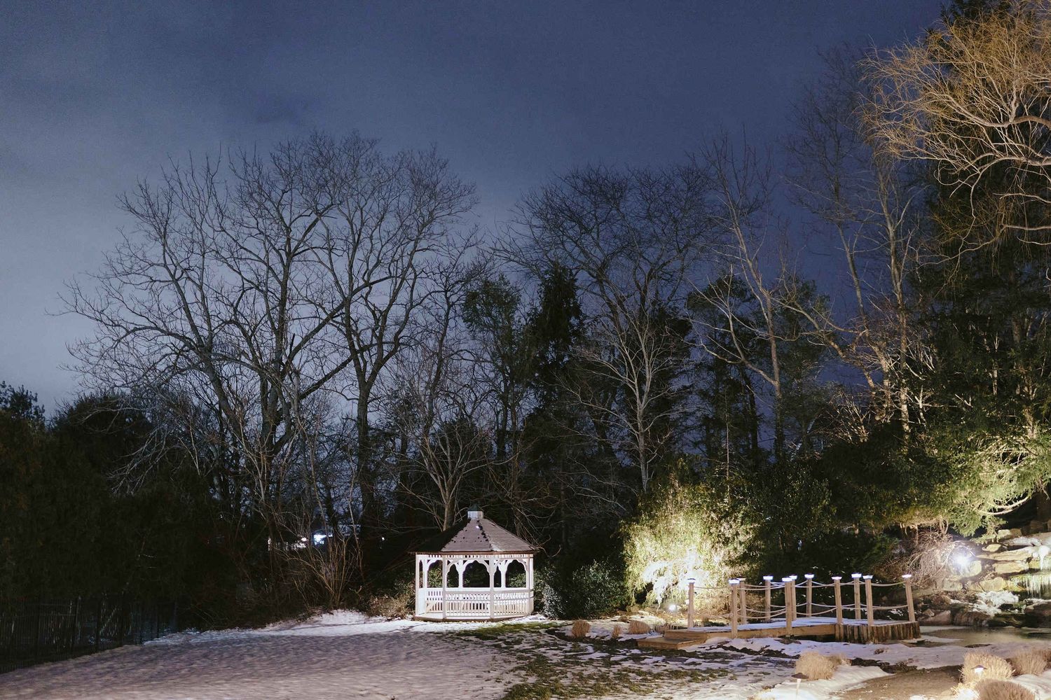 A white gazebo and buildings illuminated at night in a snowy winter landscape with bare trees.