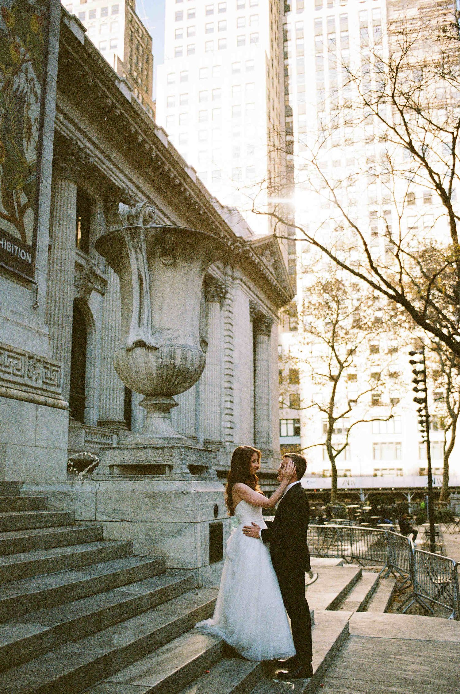 A wedding couple embraces on stone steps outside a grand architectural building in New York City.