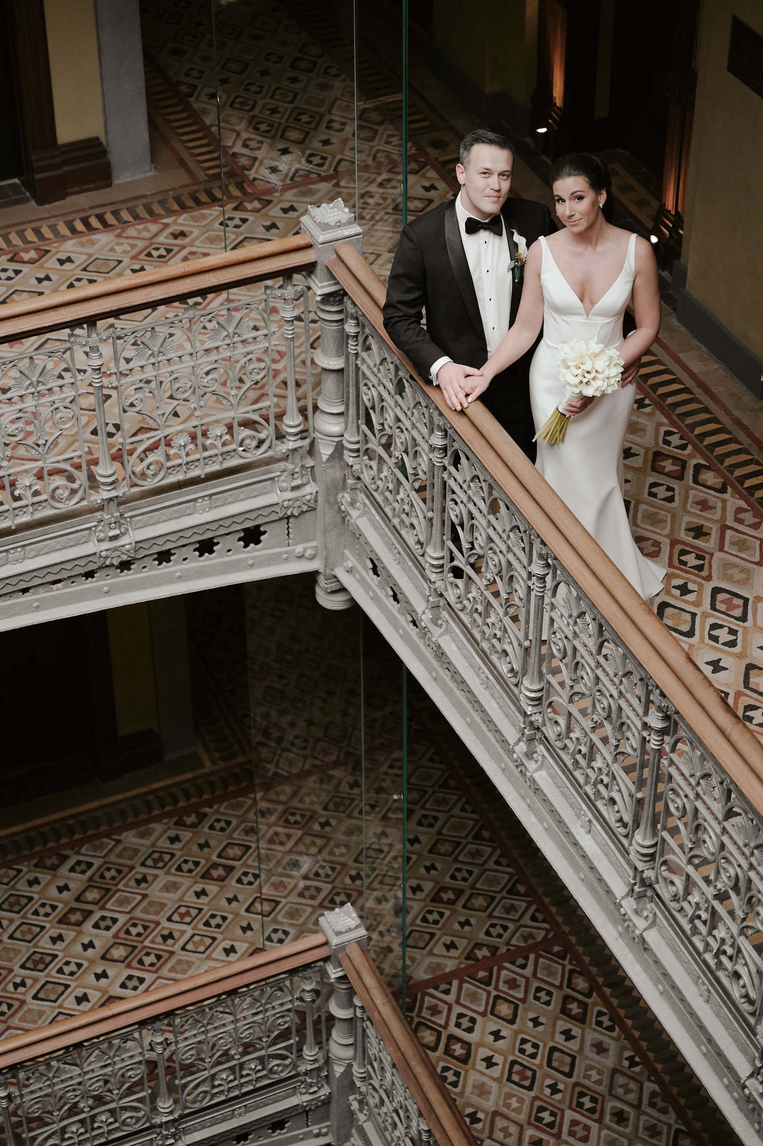 Elegant couple stands on ornate vintage staircase with intricate iron railings and patterned tile floor in historic building.