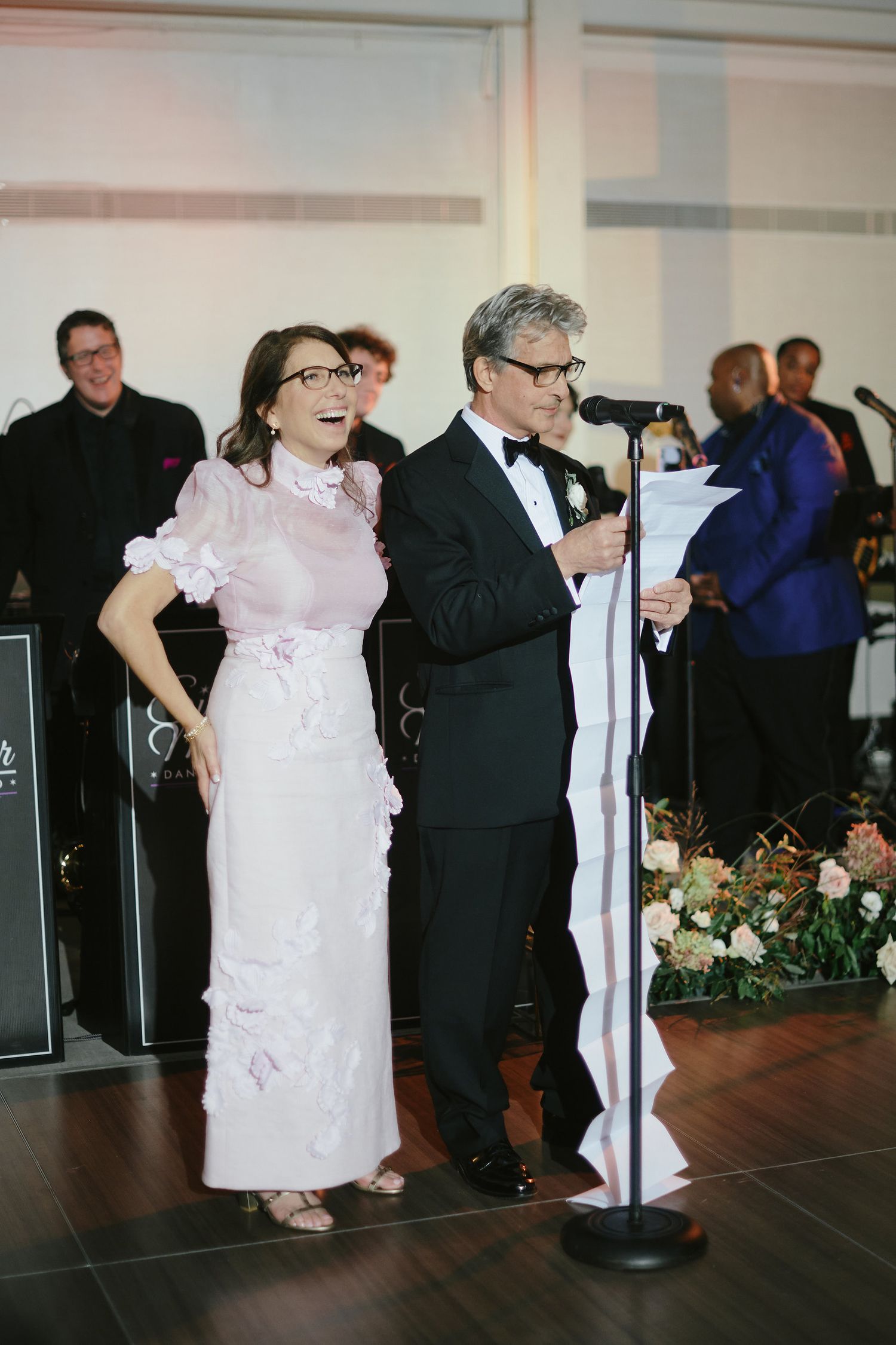 Two performers in formal attire reading from a comically long scroll at a special event with floral decorations.