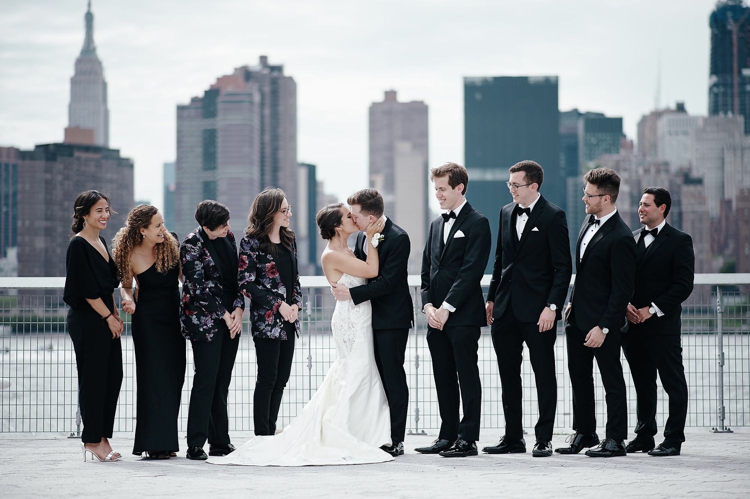 Wedding party poses together against New York City skyline including Empire State Building on waterfront.
