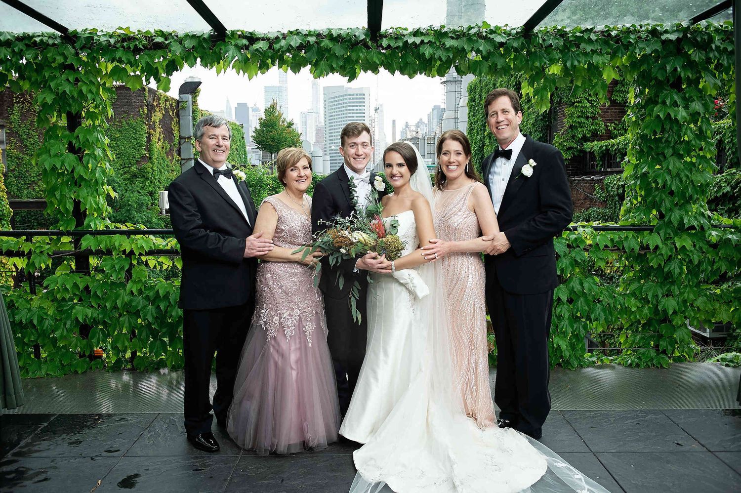A wedding party poses together under a lush green vine-covered pergola on a stone patio.