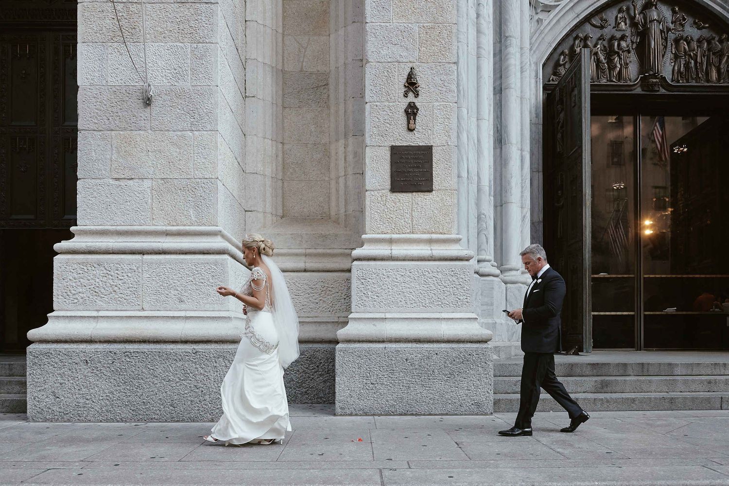 A couple in formal wedding attire playfully pose between architectural columns of a grand stone building.