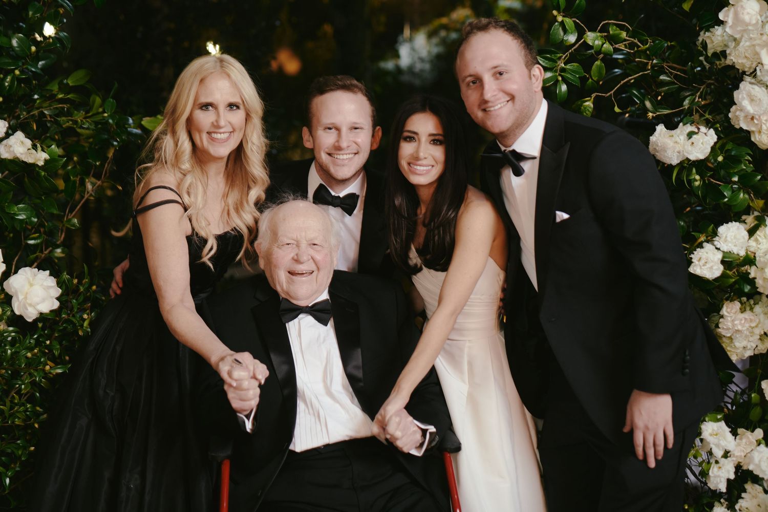A group in formal attire poses together among white flowers at an elegant outdoor event.