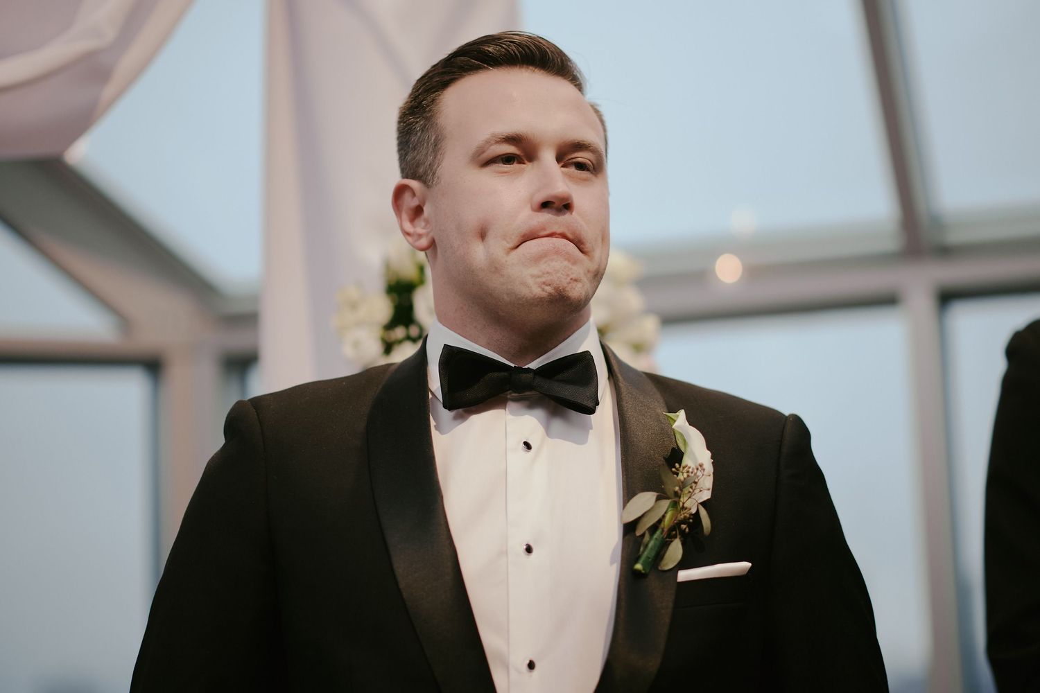 Person wearing a classic black tuxedo and bowtie with a boutonniere against a bright indoor background.