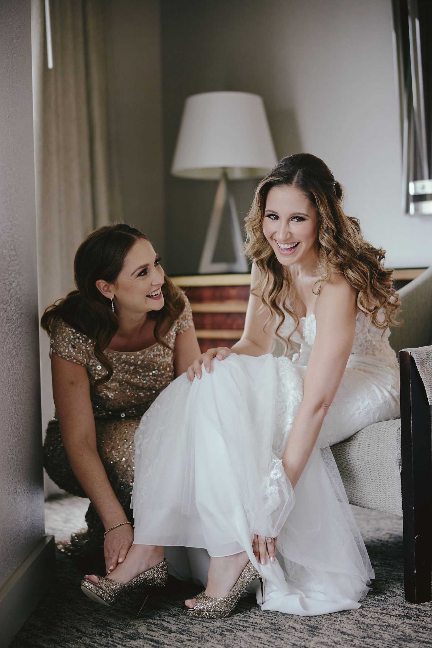 Wedding day preparations show bride in white gown and bridesmaid in sparkly dress helping with shoes on grey carpet.