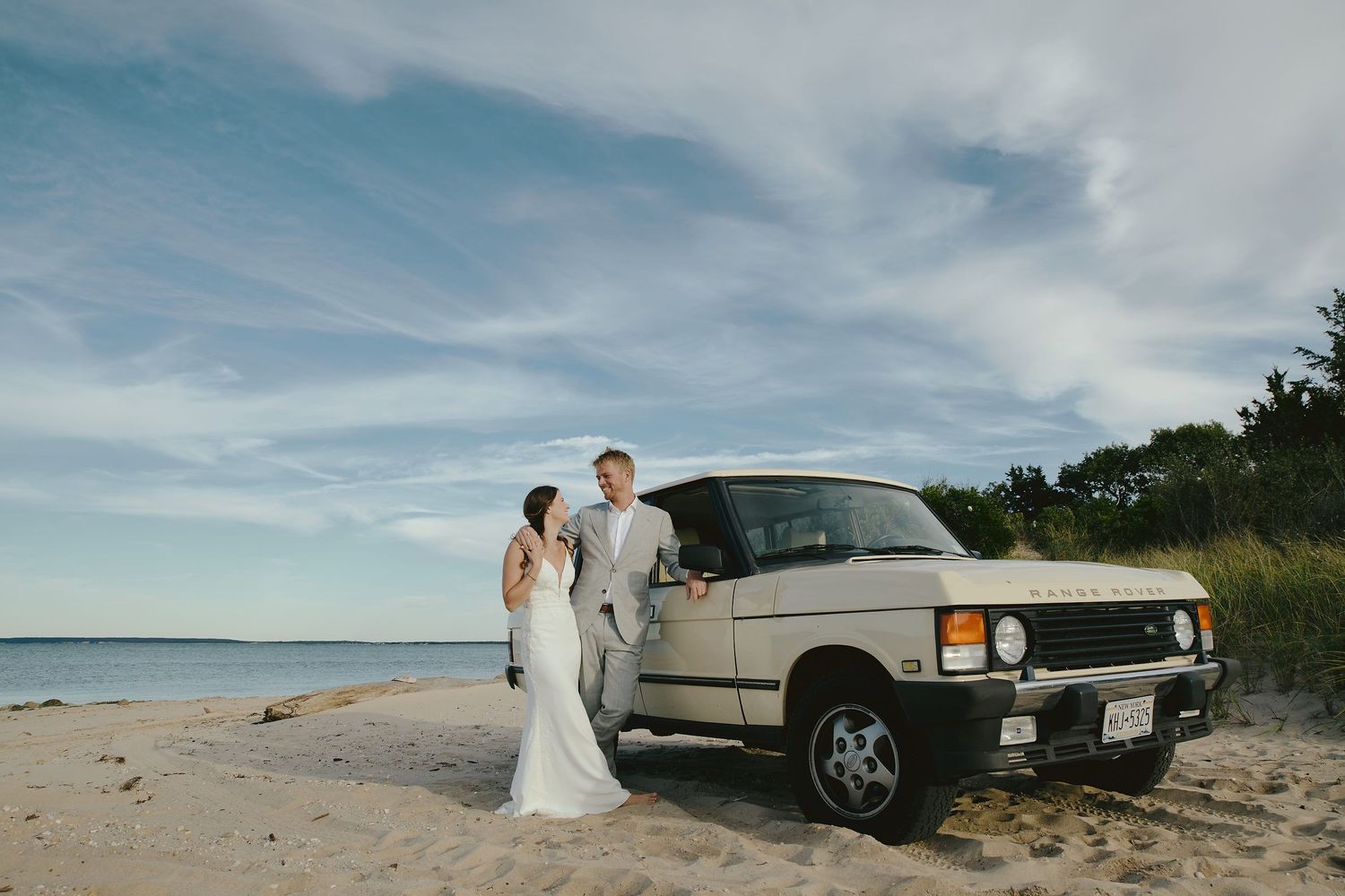 A classic Range Rover parked on a sandy beach with a scenic ocean view and cloudy sky in the background.