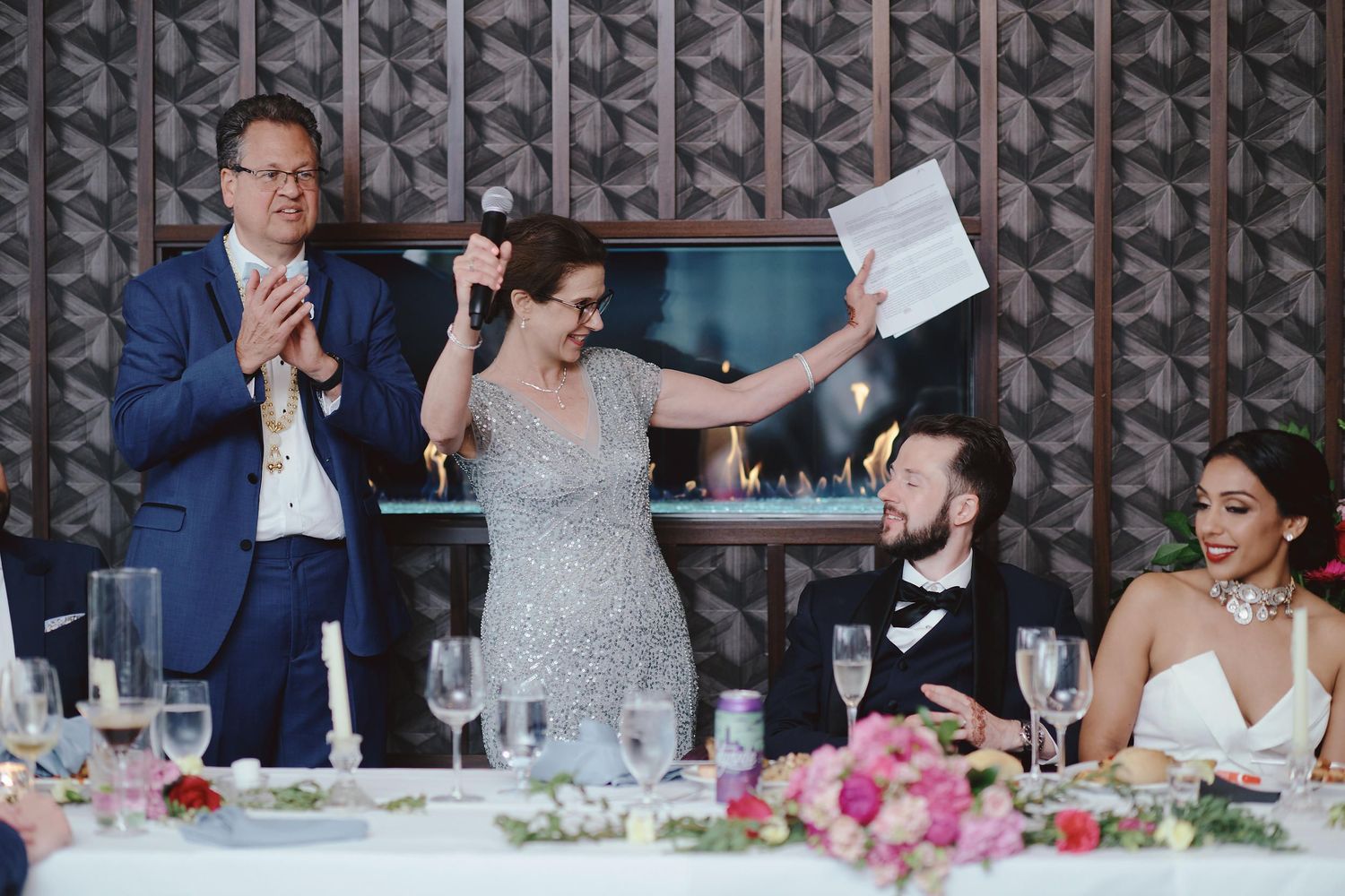 Wedding reception guests sharing joyful reactions at a decorated dining table with pink floral centerpieces.