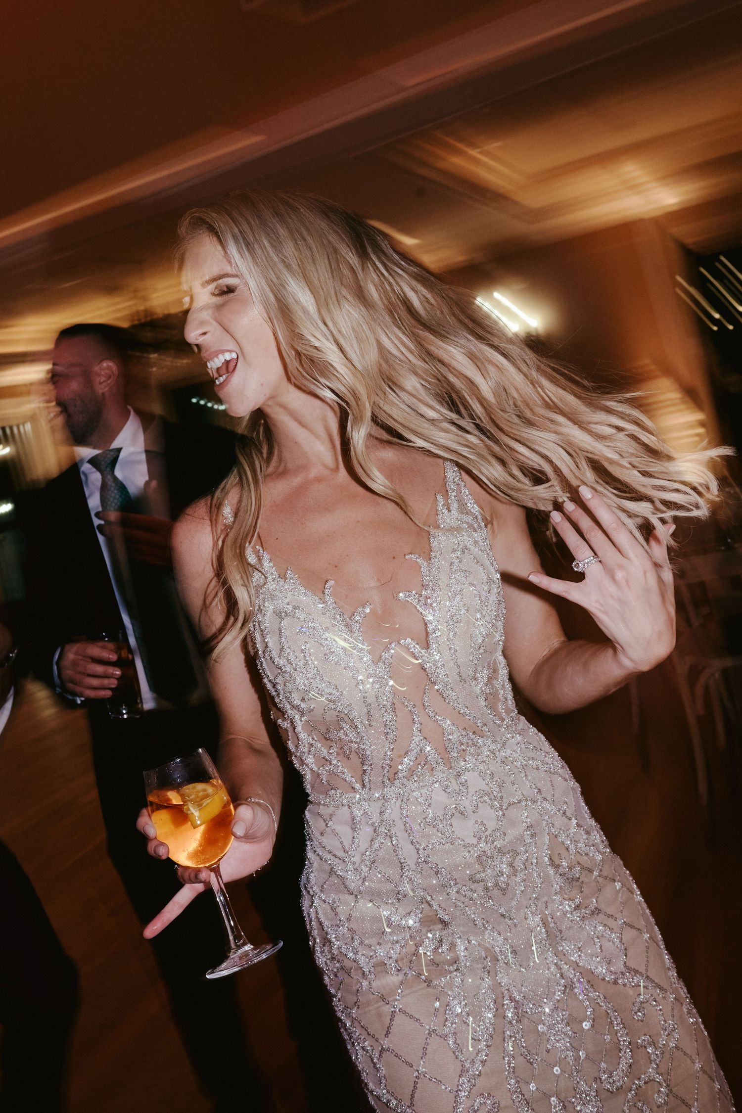 Joyful guest in sparkly lace wedding dress dancing with cocktail in hand at reception party.