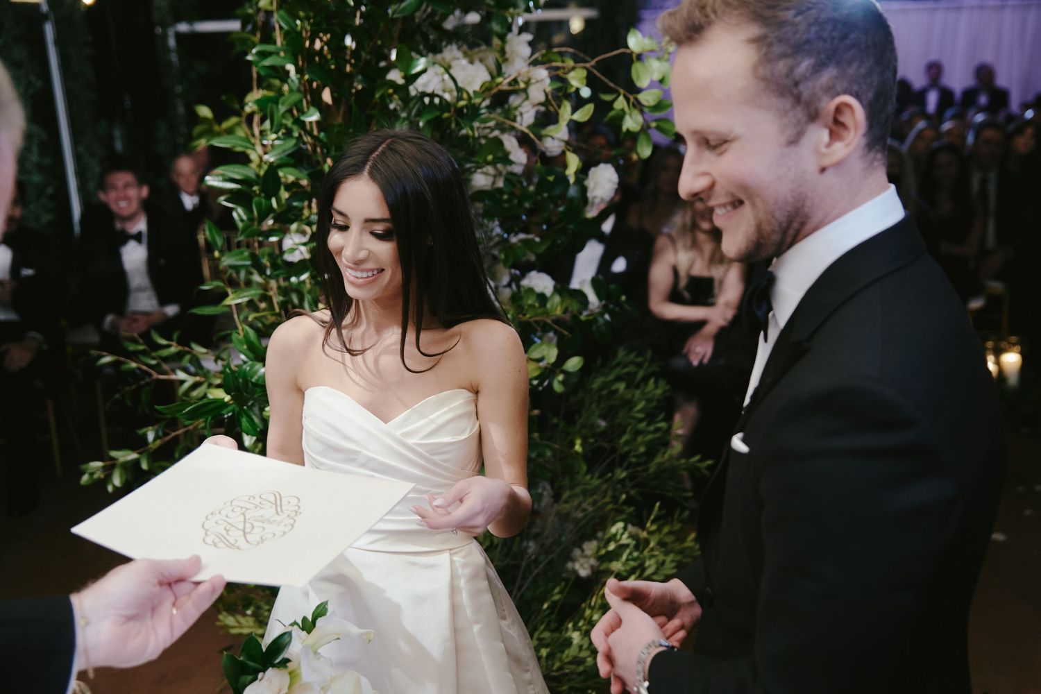 A bride and groom share a laugh while looking at paper during an elegant evening wedding ceremony with greenery decor.