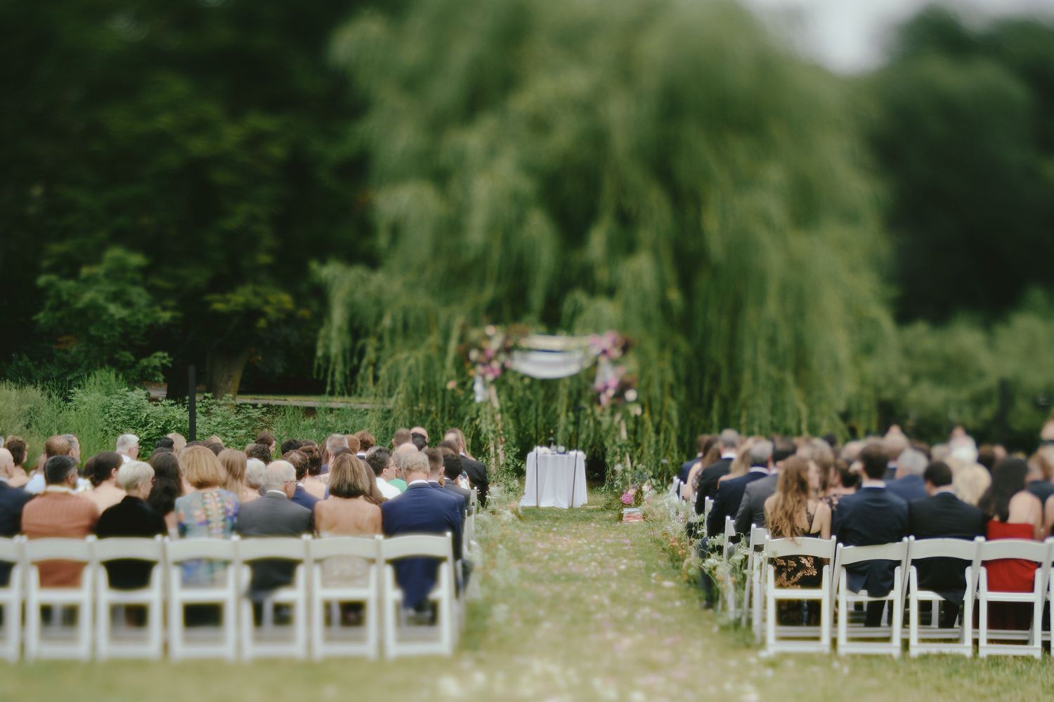 An outdoor wedding ceremony setup with white chairs and a floral arch under a weeping willow tree.