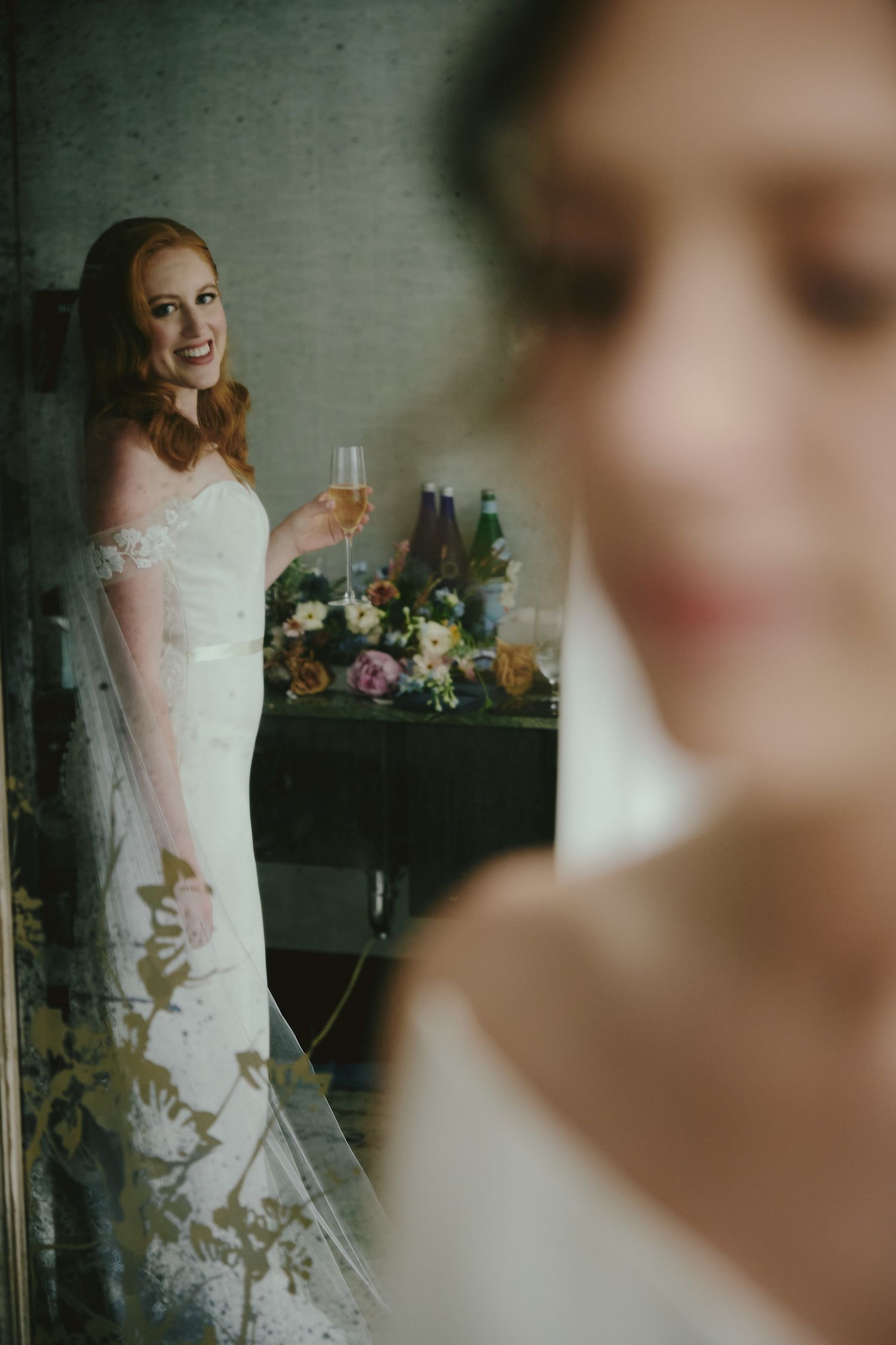 A bride in an elegant white lace gown poses with a champagne glass against a wall with floral decorations.